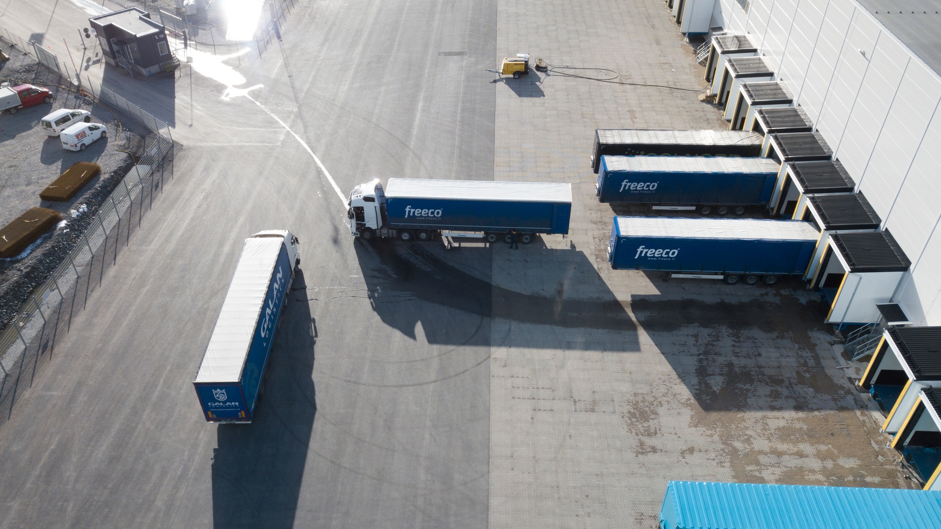 Flygfoto över ett logistikcentrum med flera blå lastbilar parkerade på en betongyta tillsammans med lastningsplattformar och containrar.