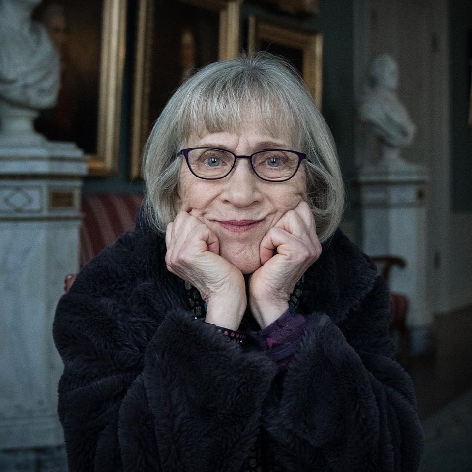 Porträtt av Claudia Goldin med kort grått hår och glasögon som ler vänligt mot kameran med händerna framför ansiktet. Hon bär en mörk kläder och fotograferas i en elegant inomhusmiljö med klassiska skulpturer och inramade konstverk synliga i bakgrunden.