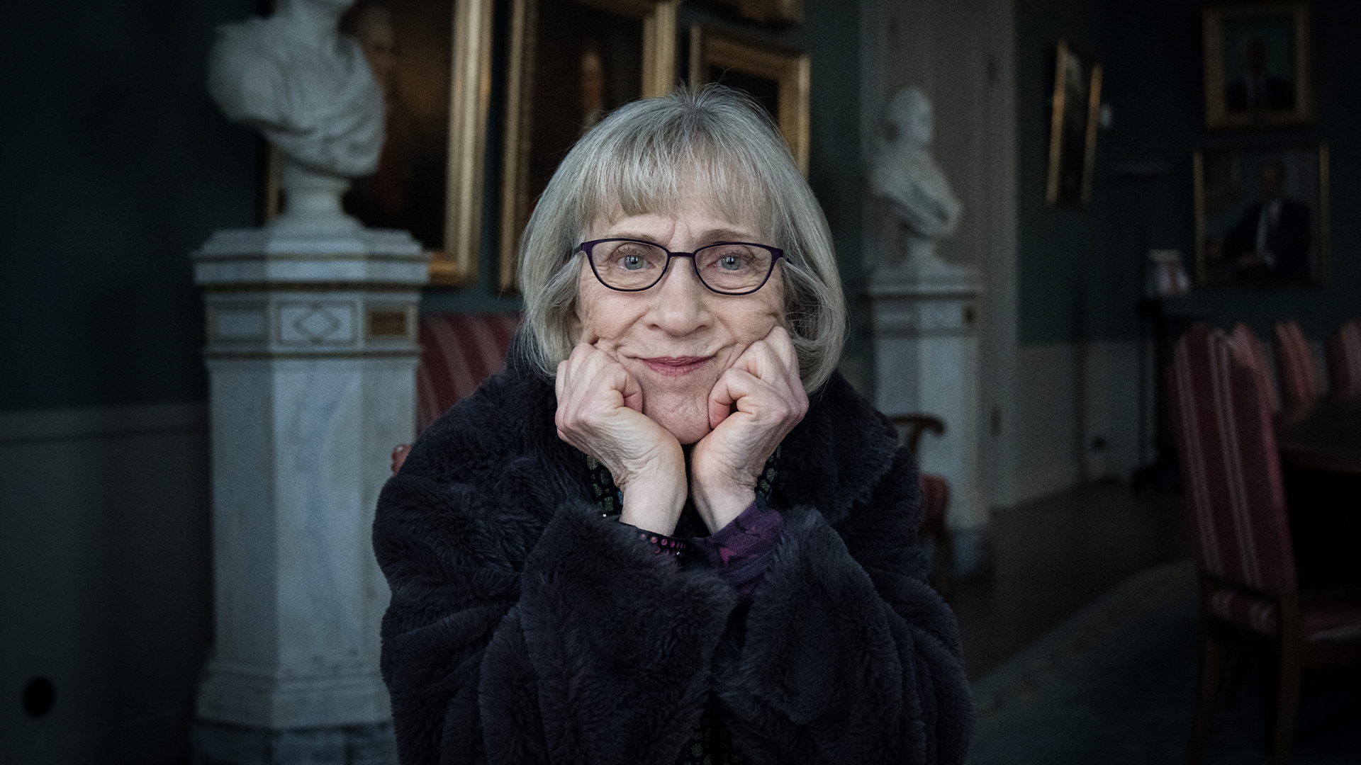 Porträtt av Claudia Goldin med kort grått hår och glasögon som ler vänligt mot kameran med händerna framför ansiktet. Hon bär en mörk kläder och fotograferas i en elegant inomhusmiljö med klassiska skulpturer och inramade konstverk synliga i bakgrunden.