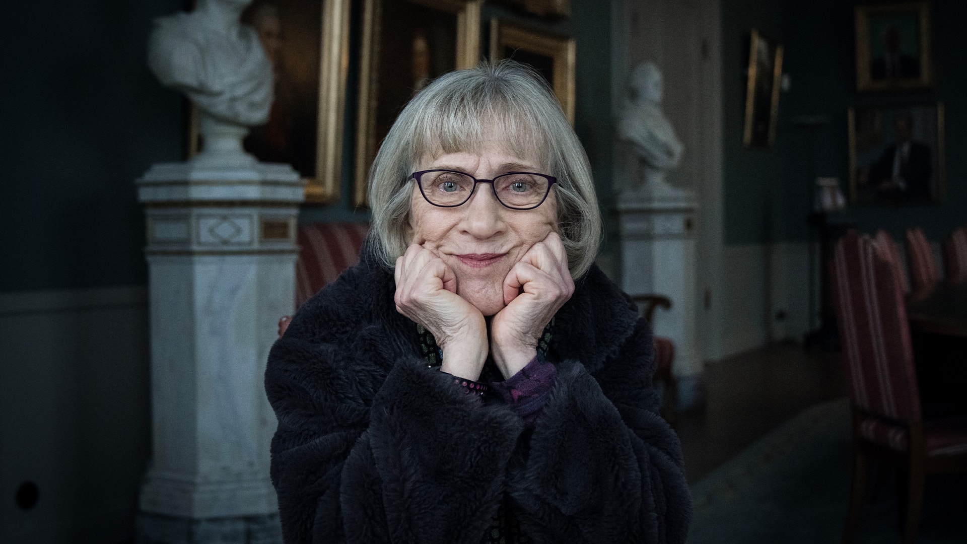 Porträtt av Claudia Goldin med kort grått hår och glasögon som ler vänligt mot kameran med händerna framför ansiktet. Hon bär en mörk kläder och fotograferas i en elegant inomhusmiljö med klassiska skulpturer och inramade konstverk synliga i bakgrunden.