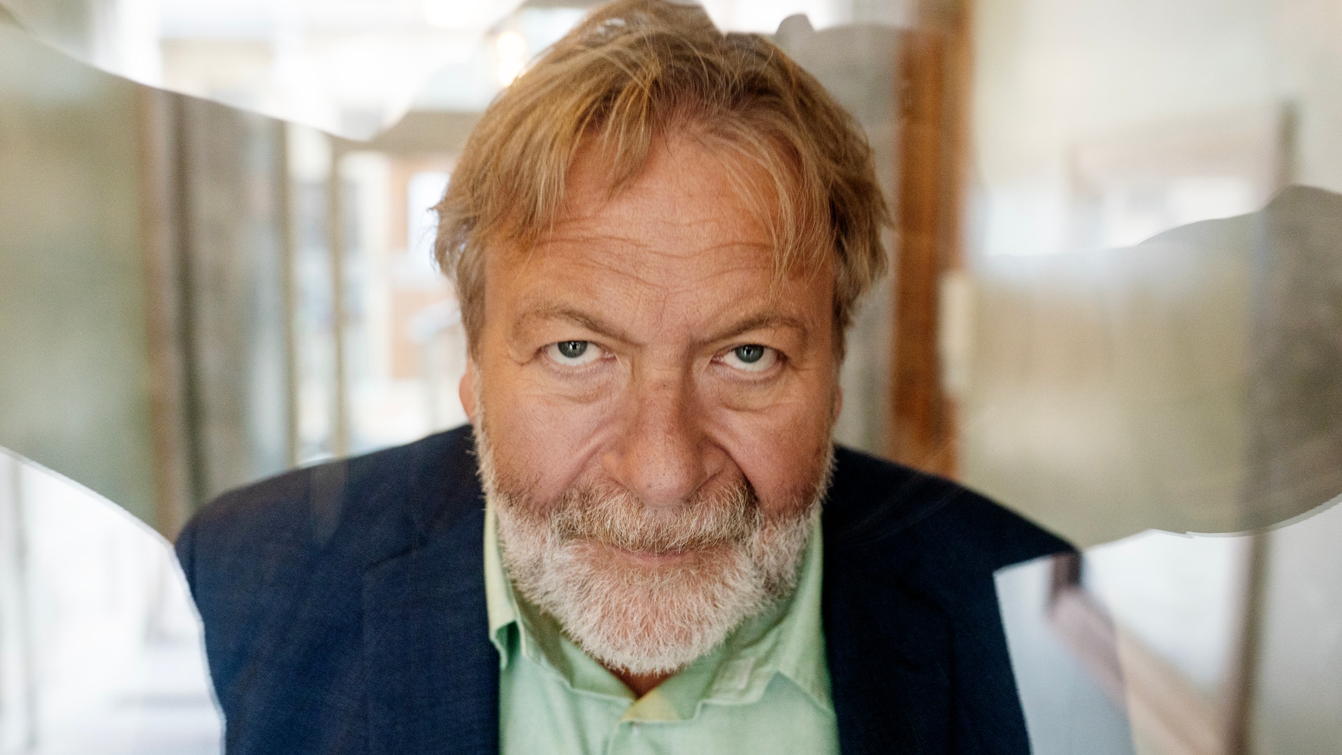 Närbild på en äldre man med grått skägg som tittar åt sidan, fotograferad genom ett fönster.