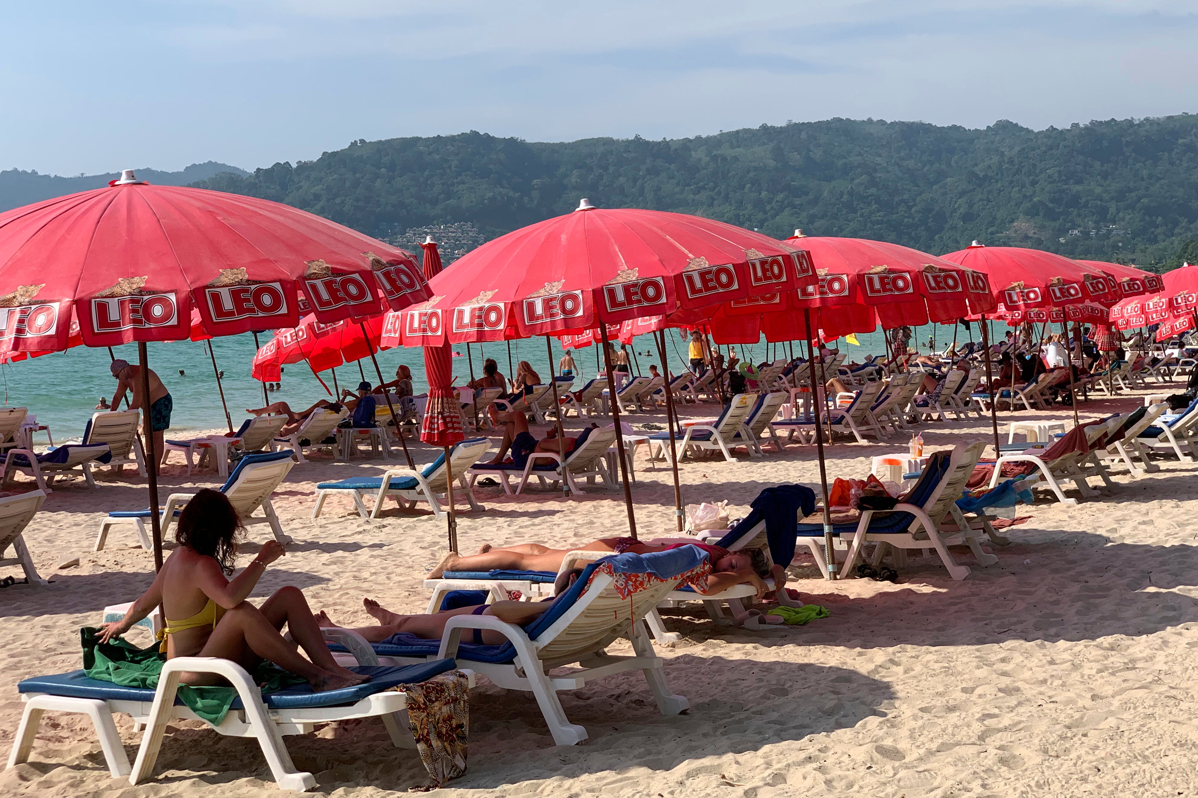 En strand med vita solstolar under röda parasoller. Människor vilar på stranden med grön skog och berg i bakgrunden.