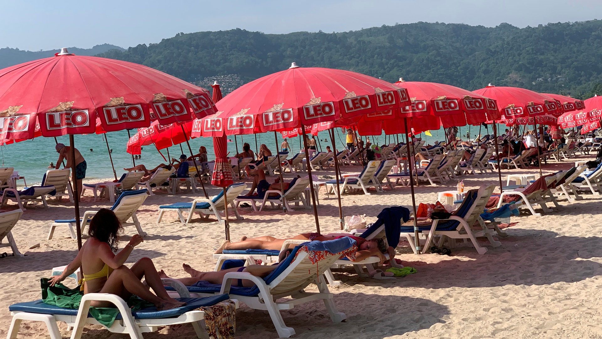 En strand med vita solstolar under röda parasoller. Människor vilar på stranden med grön skog och berg i bakgrunden.