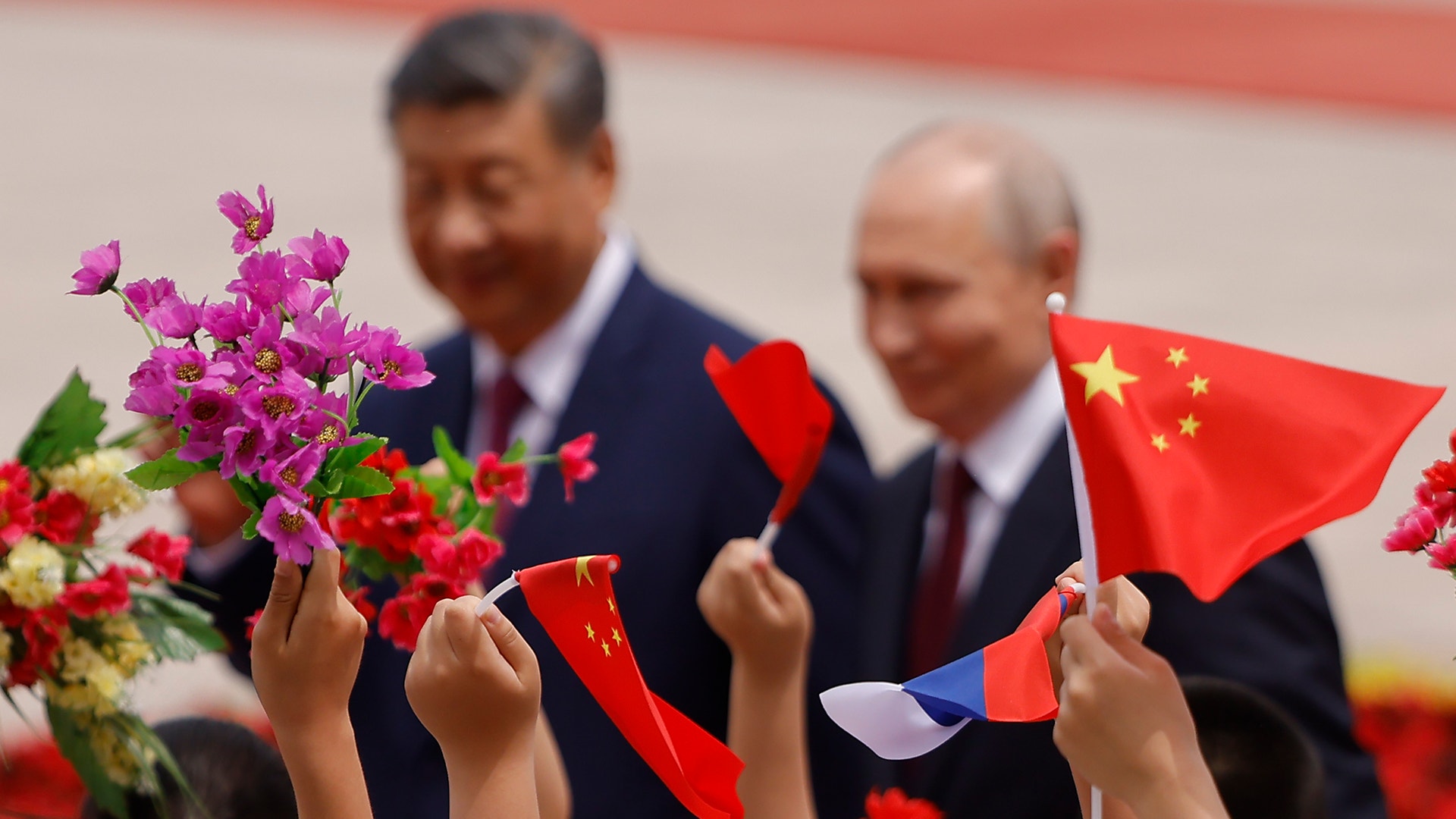 Xi Jinping och Vladimir Putin i bakgrunden. I förgrunden håller händer upp kinesiska flaggor och rosa och röda blomster.