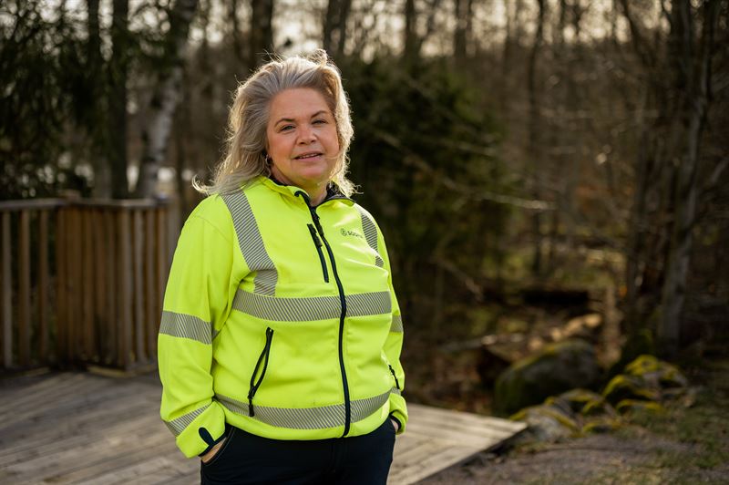 Porträtt av en person i gul reflexväst som står på en trädäck med skog i bakgrunden.