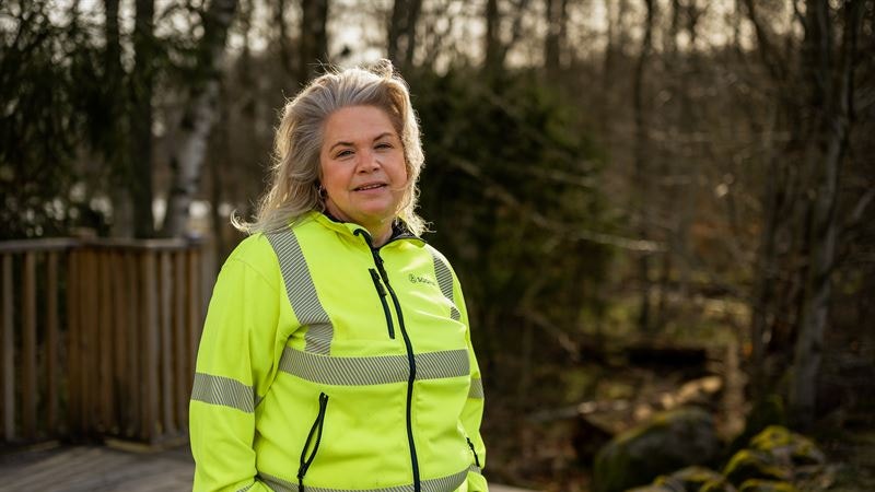 Porträtt av en person i gul reflexväst som står på en trädäck med skog i bakgrunden.