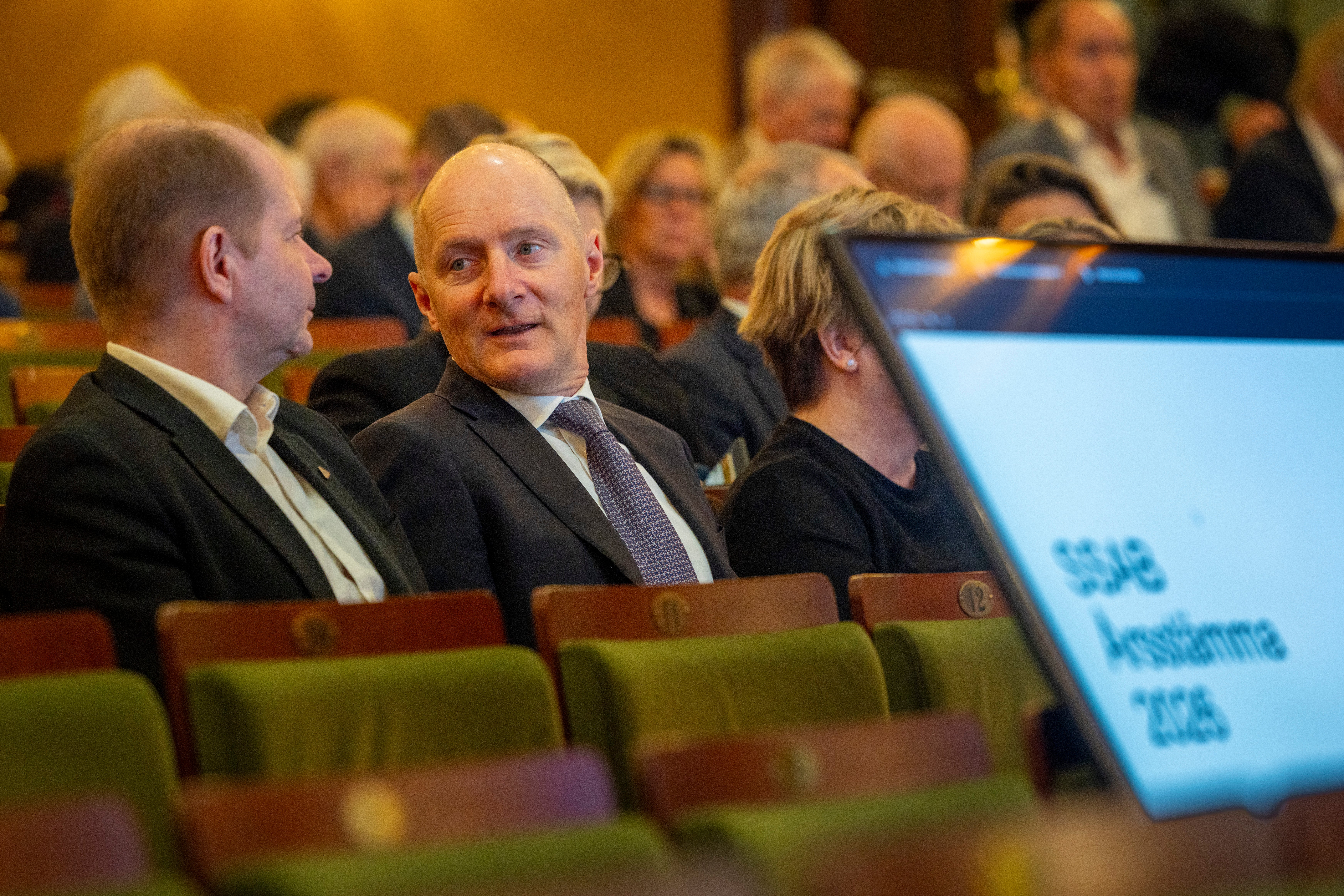 Män i kostym sitter på gröna stolar under ett möte eller en konferens i en auditorieum.