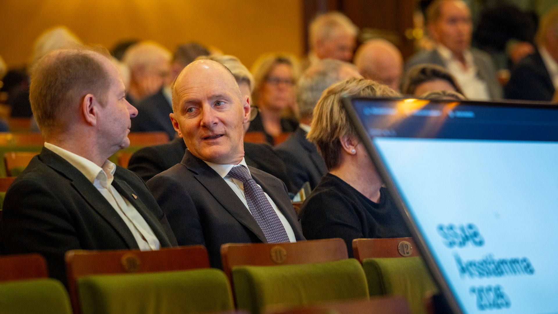 Män i kostym sitter på gröna stolar under ett möte eller en konferens i en auditorieum.