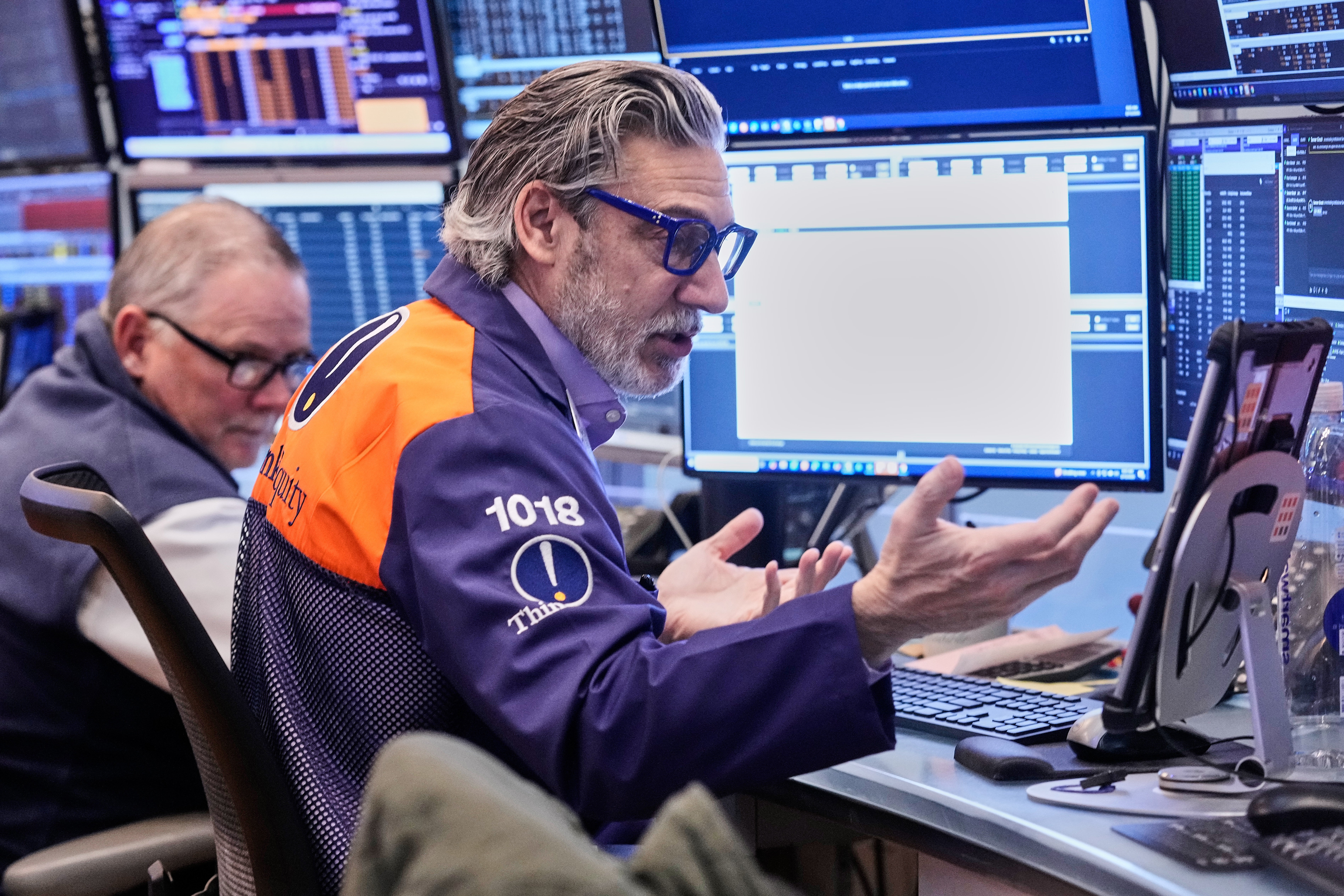 Två aktiehandlare arbetar vid sina arbetsplatser på New York Stock Exchange. Mannen i förgrunden bär en orange och mörkblå väst och glasögon, och gestikulerar medan han följer marknadsdata på flera datorskärmar som visar realtidsaktiekurser och finansiell information. En annan handlare sitter bakom honom. Bakgrunden visar ytterligare skärmar med finansiell data och handelsgränssnitt.