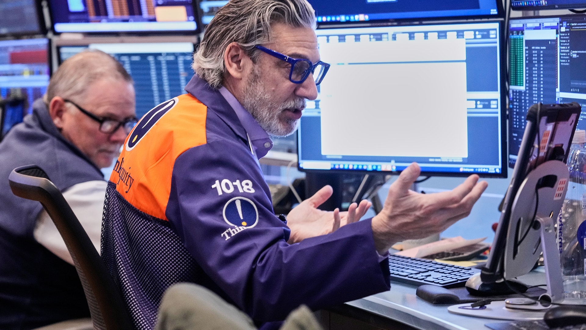 Två aktiehandlare arbetar vid sina arbetsplatser på New York Stock Exchange. Mannen i förgrunden bär en orange och mörkblå väst och glasögon, och gestikulerar medan han följer marknadsdata på flera datorskärmar som visar realtidsaktiekurser och finansiell information. En annan handlare sitter bakom honom. Bakgrunden visar ytterligare skärmar med finansiell data och handelsgränssnitt.