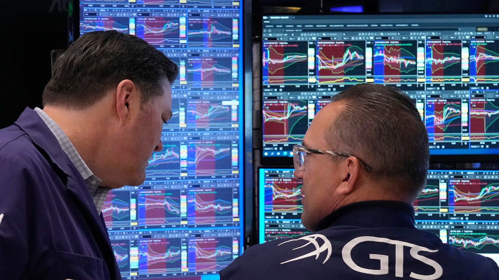 Två män tittar på finansiella handelsskärmar med grafer och marknadsdata.