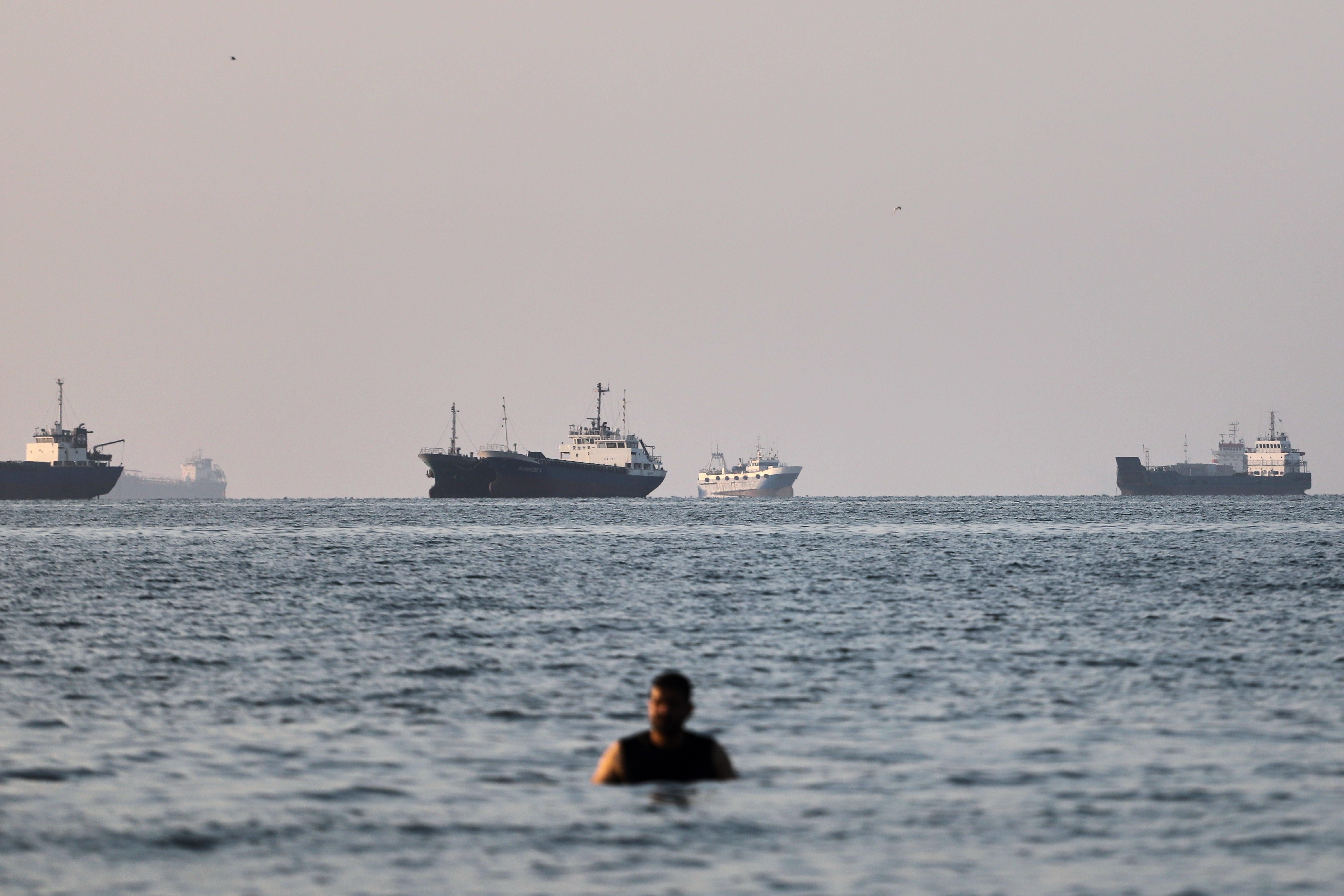 En person badar i havet medan flera lastfartyg ligger ankrade på horisonten i bakgrunden.