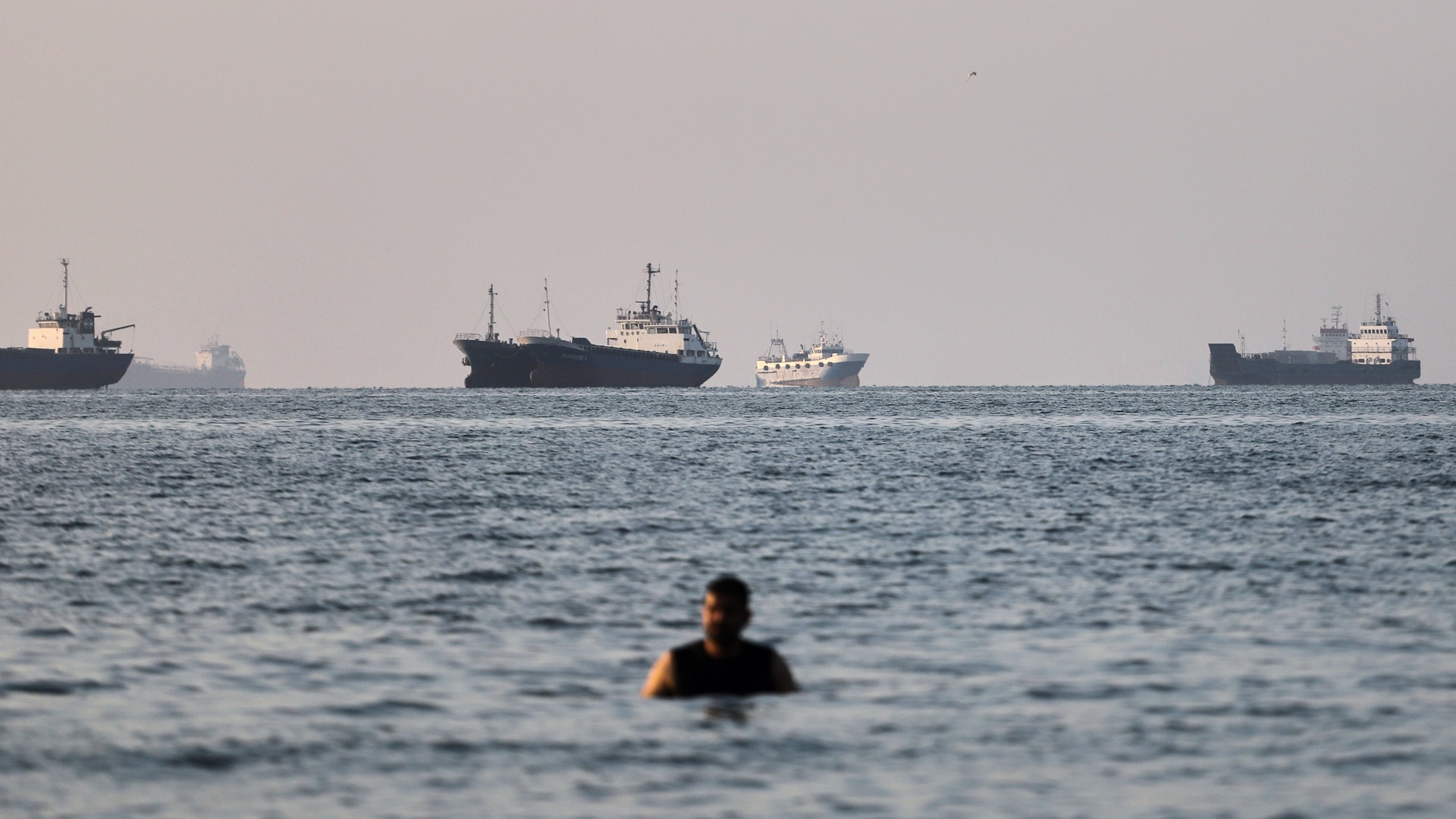 En person badar i havet medan flera lastfartyg ligger ankrade på horisonten i bakgrunden.