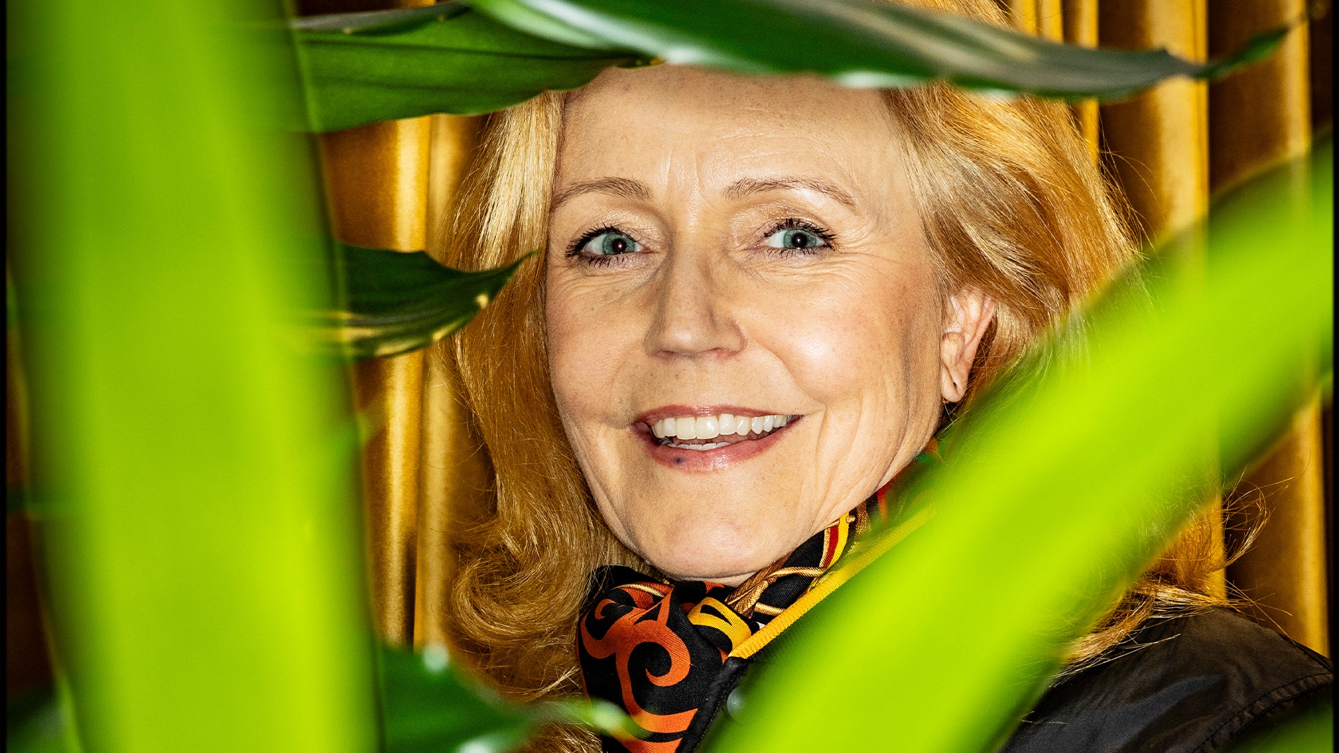 Porträtt av en leende kvinna med rödblont hår, iklädd mörk jacka och mönstrad scarf, inramad av gröna växtblad.