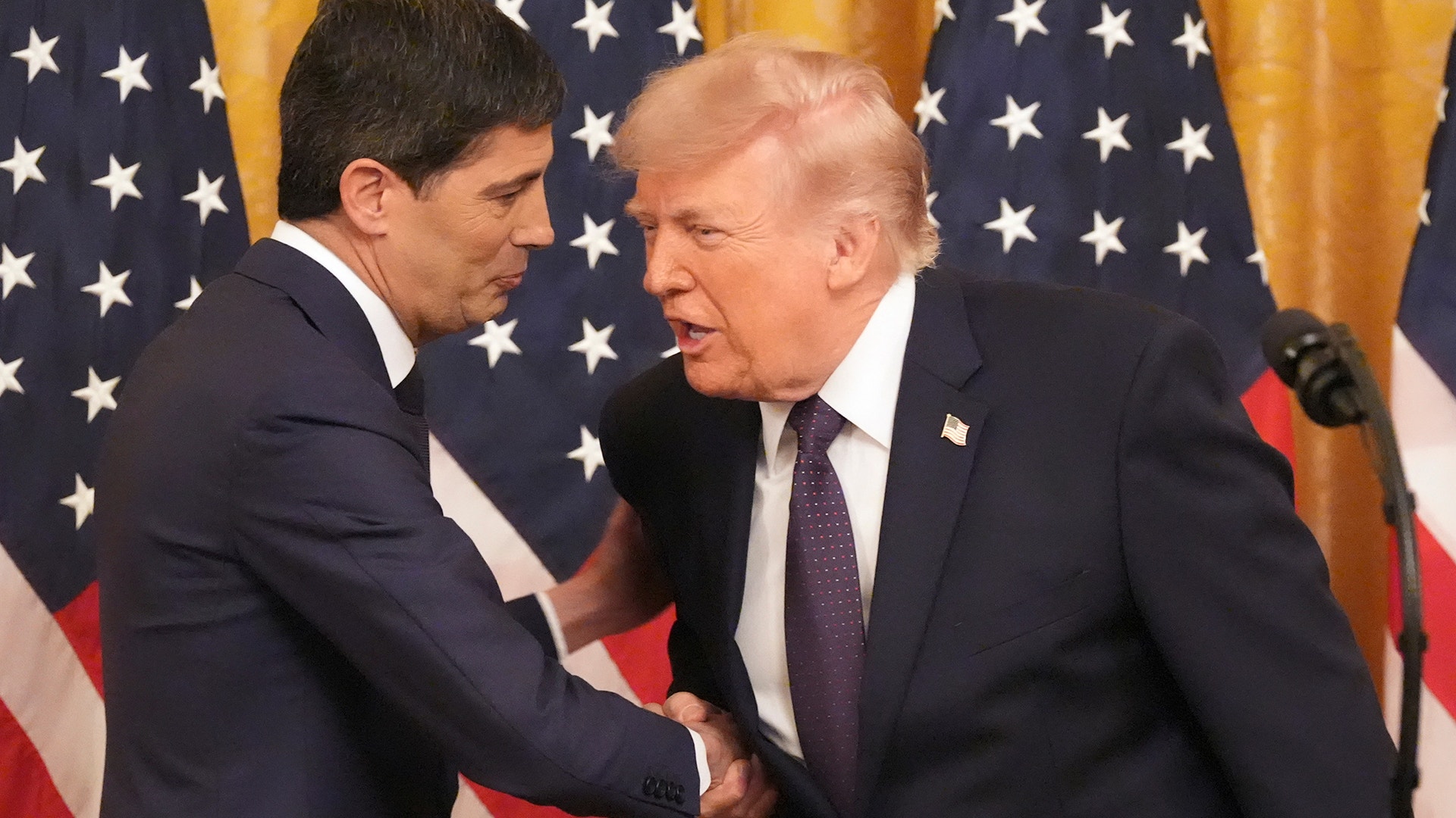 Två män i kostym skakar hand framför amerikanska flaggor och guldgardiner vid ett officiellt möte.