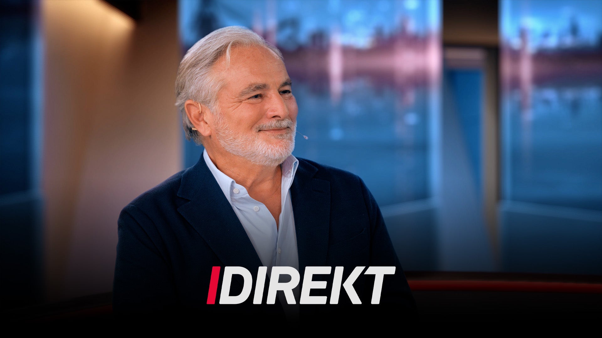 En äldre man med vitt hår och skägg leende i en tv-studio, med texten "Direkt" i bild.