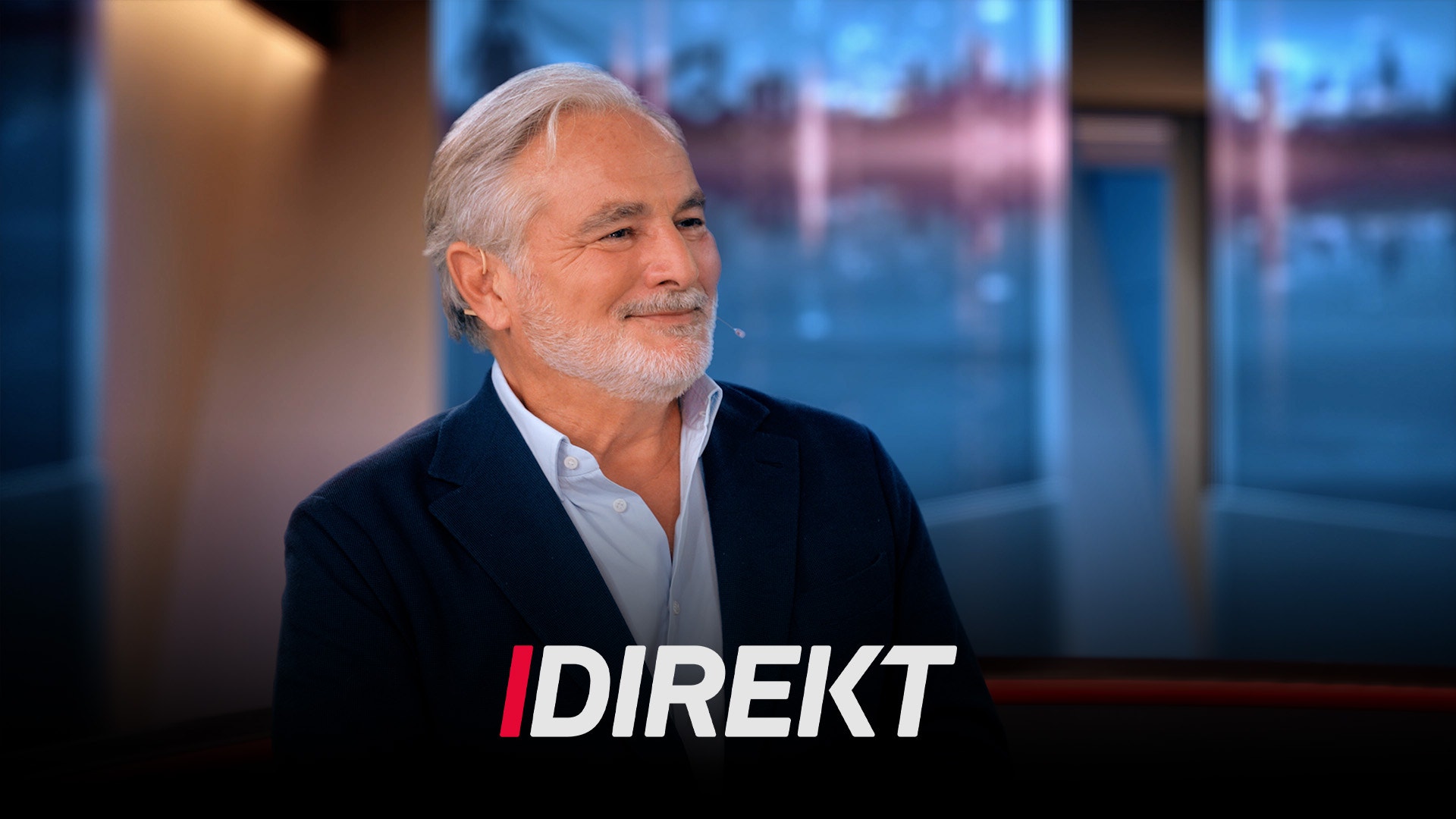 En äldre man med vitt hår och skägg leende i en tv-studio, med texten "Direkt" i bild.