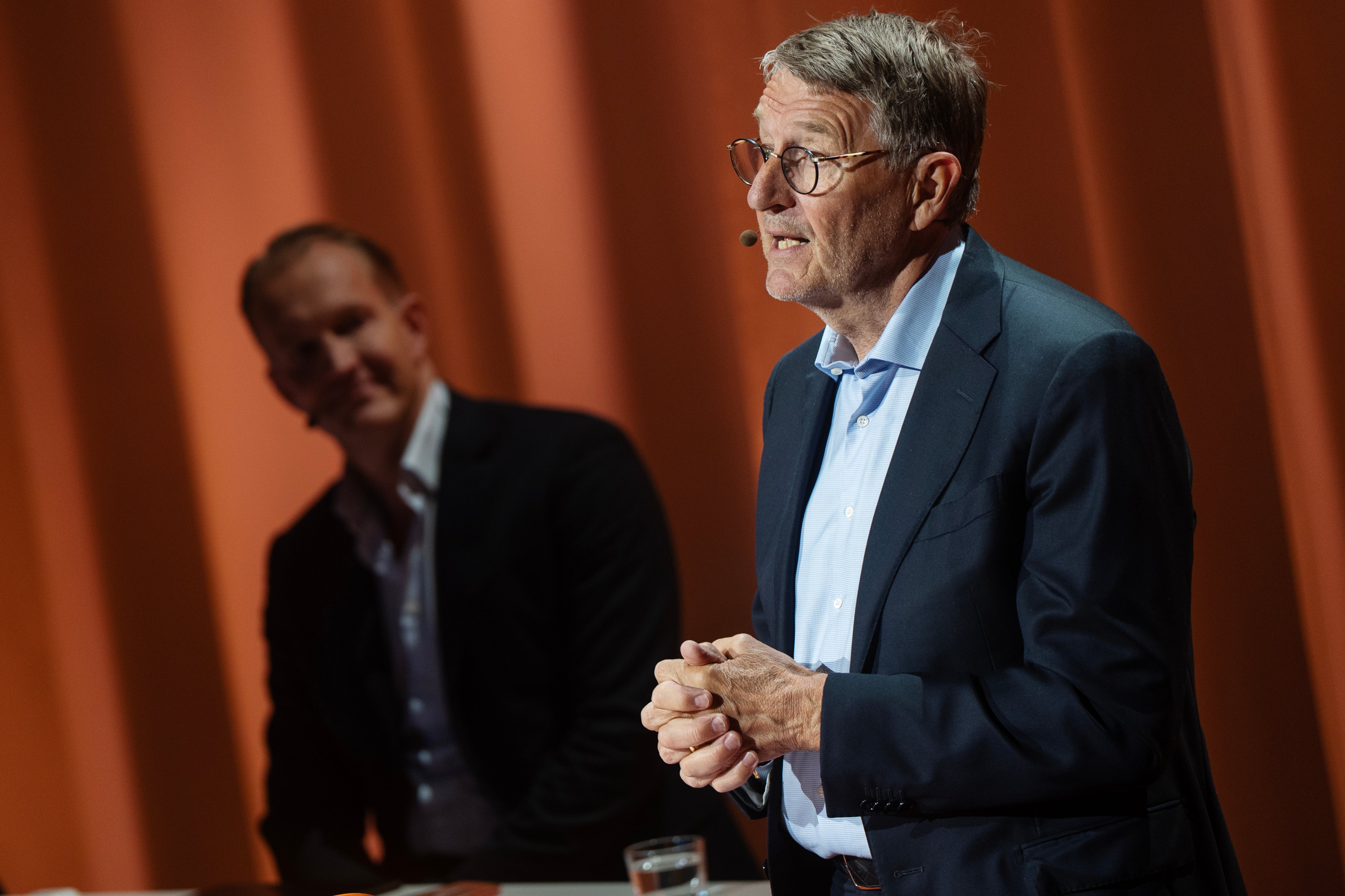 En man i blå kostym och glasögon talar vid en stämma framför en orange gardin. En annan person syns i bakgrunden.