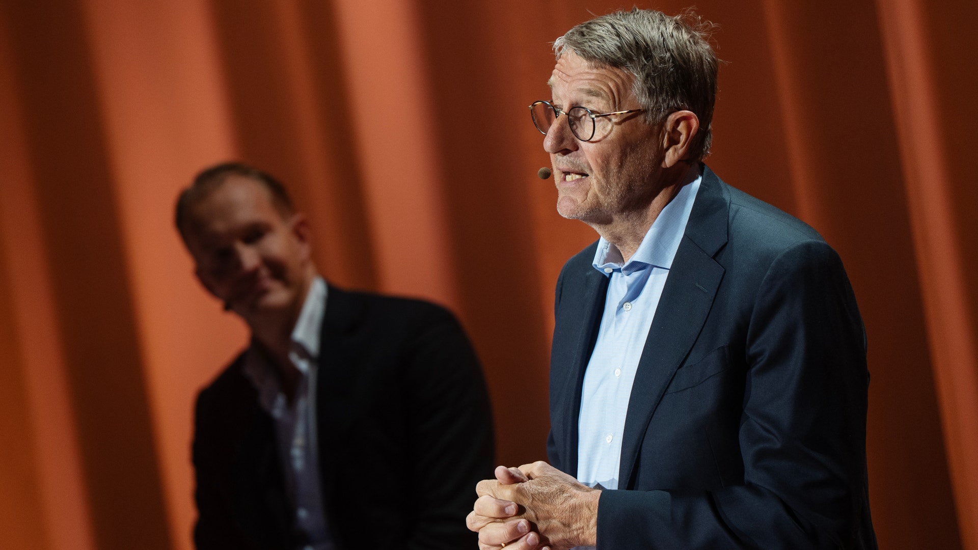 En man i blå kostym och glasögon talar vid en stämma framför en orange gardin. En annan person syns i bakgrunden.