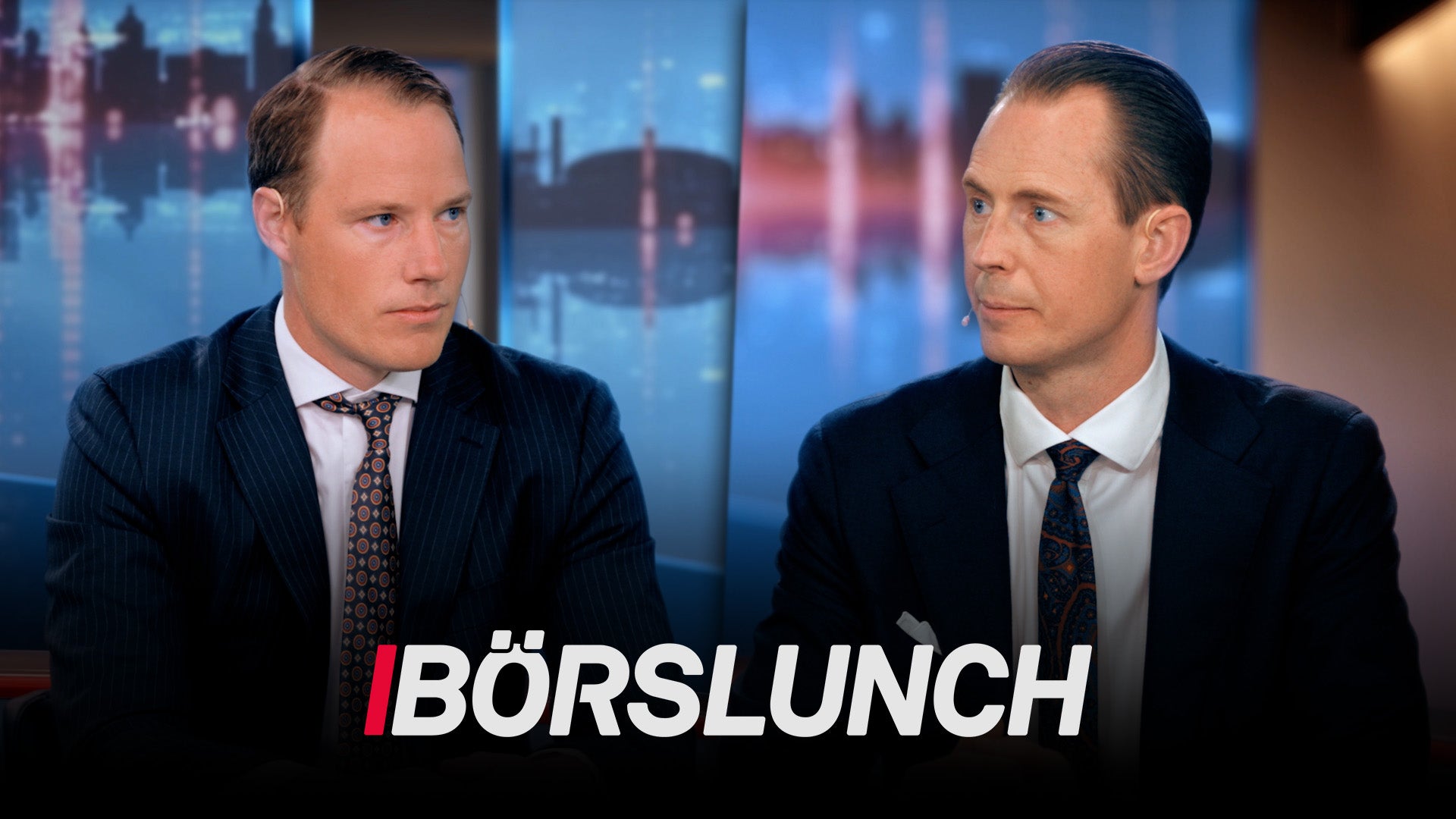 Två män i affärskläder sitter vid ett studiobord i TV-programmet Börslunch med en stadshorisont i bakgrunden.