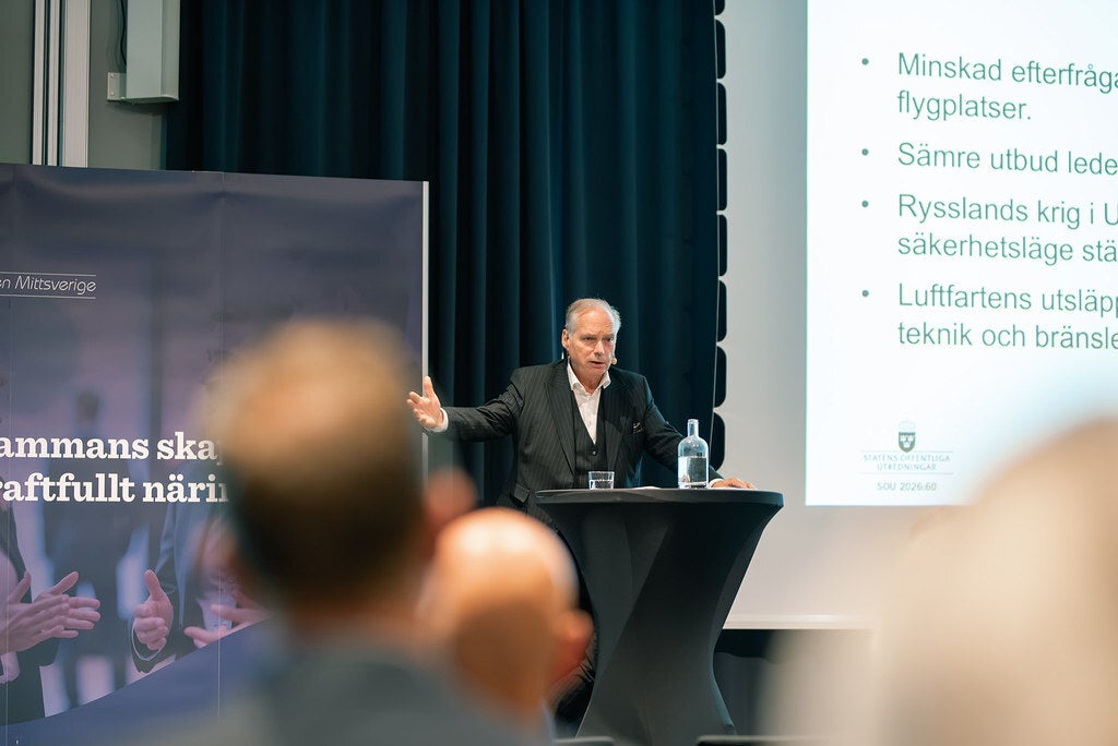 En man håller en presentation för en publik, med en bildskärm med punktlistor i bakgrunden.
