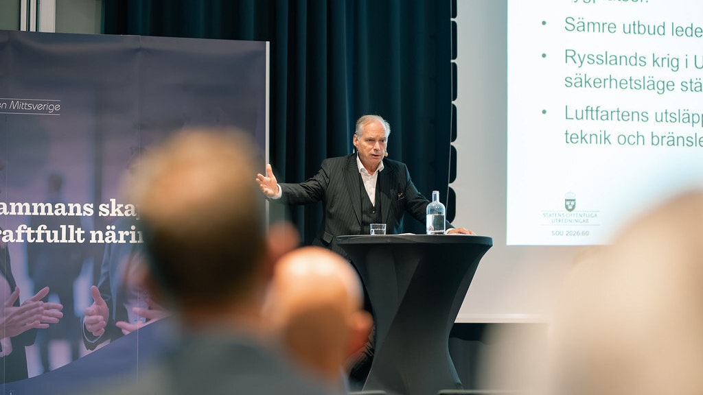 En man håller en presentation för en publik, med en bildskärm med punktlistor i bakgrunden.
