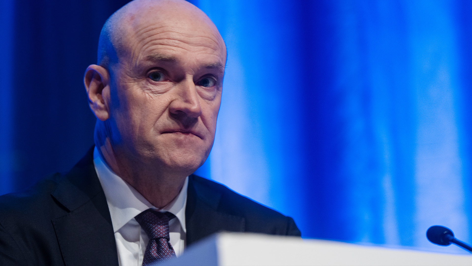 En man i kostym talar vid ett podium framför en blå bakgrund.