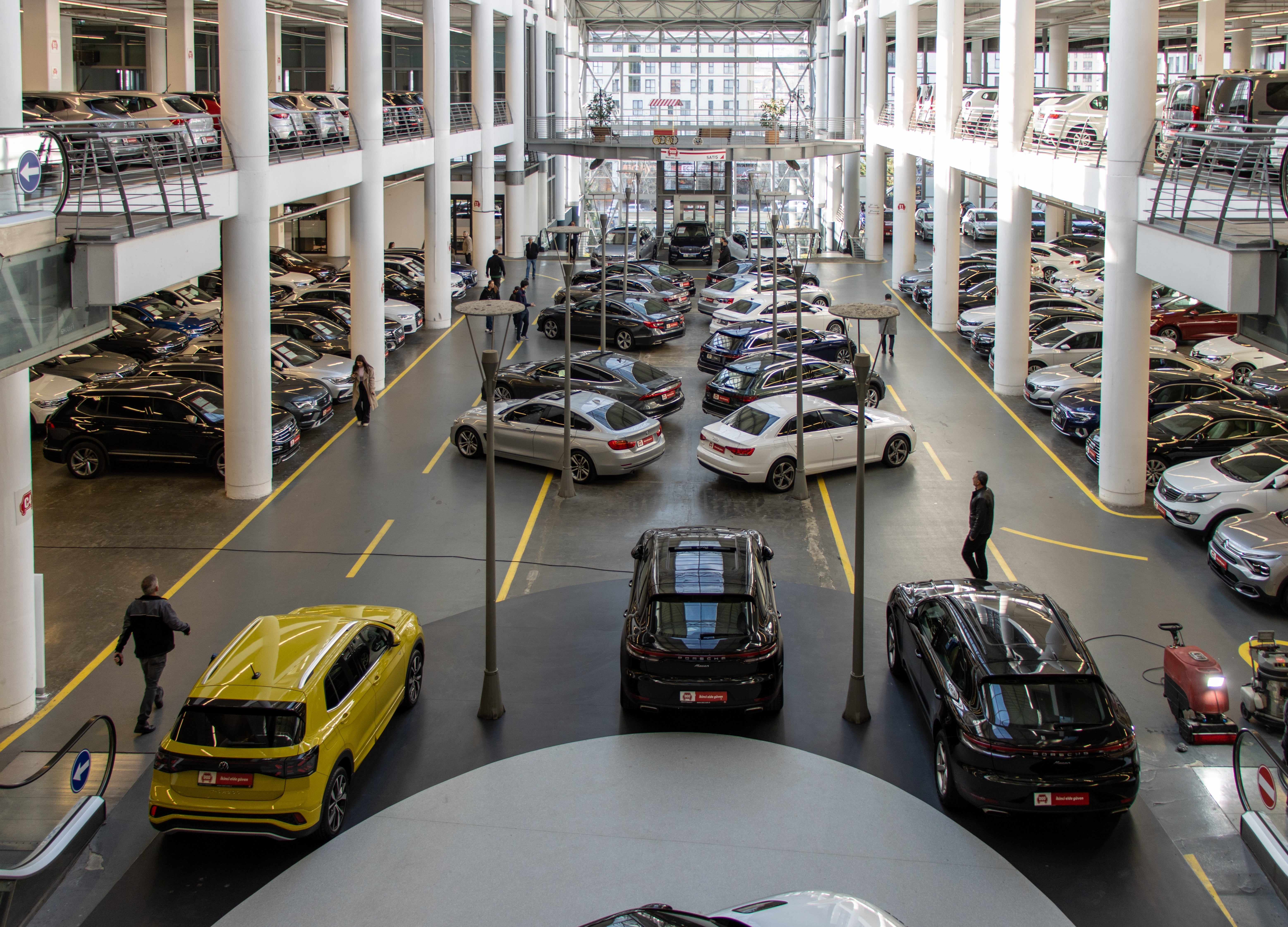 Interiör av en stor biluthyrning eller bilhandel med många bilar parkerade på flera nivåer. Vita pelare och gula parkeringsmarkeringar på golvet. En gul bil, en svart bil och andra fordon är synliga i showrummet.