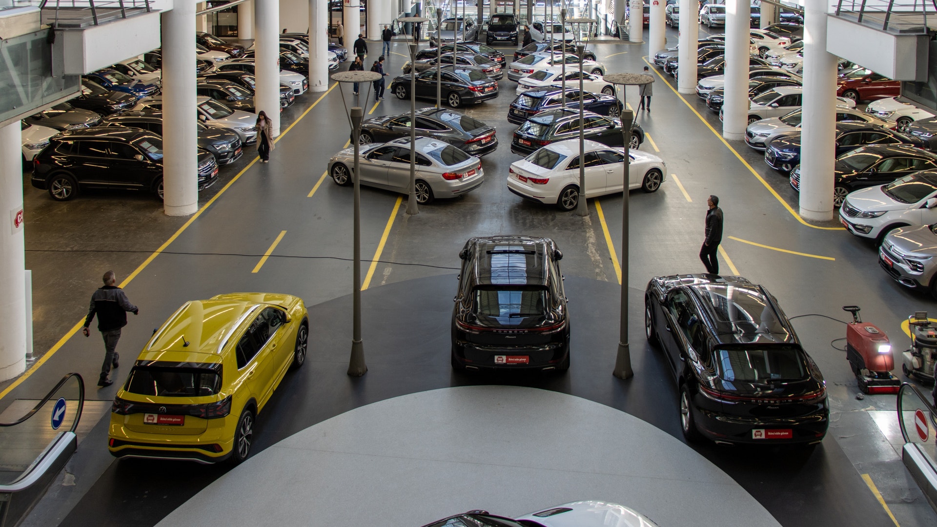Interiör av en stor biluthyrning eller bilhandel med många bilar parkerade på flera nivåer. Vita pelare och gula parkeringsmarkeringar på golvet. En gul bil, en svart bil och andra fordon är synliga i showrummet.
