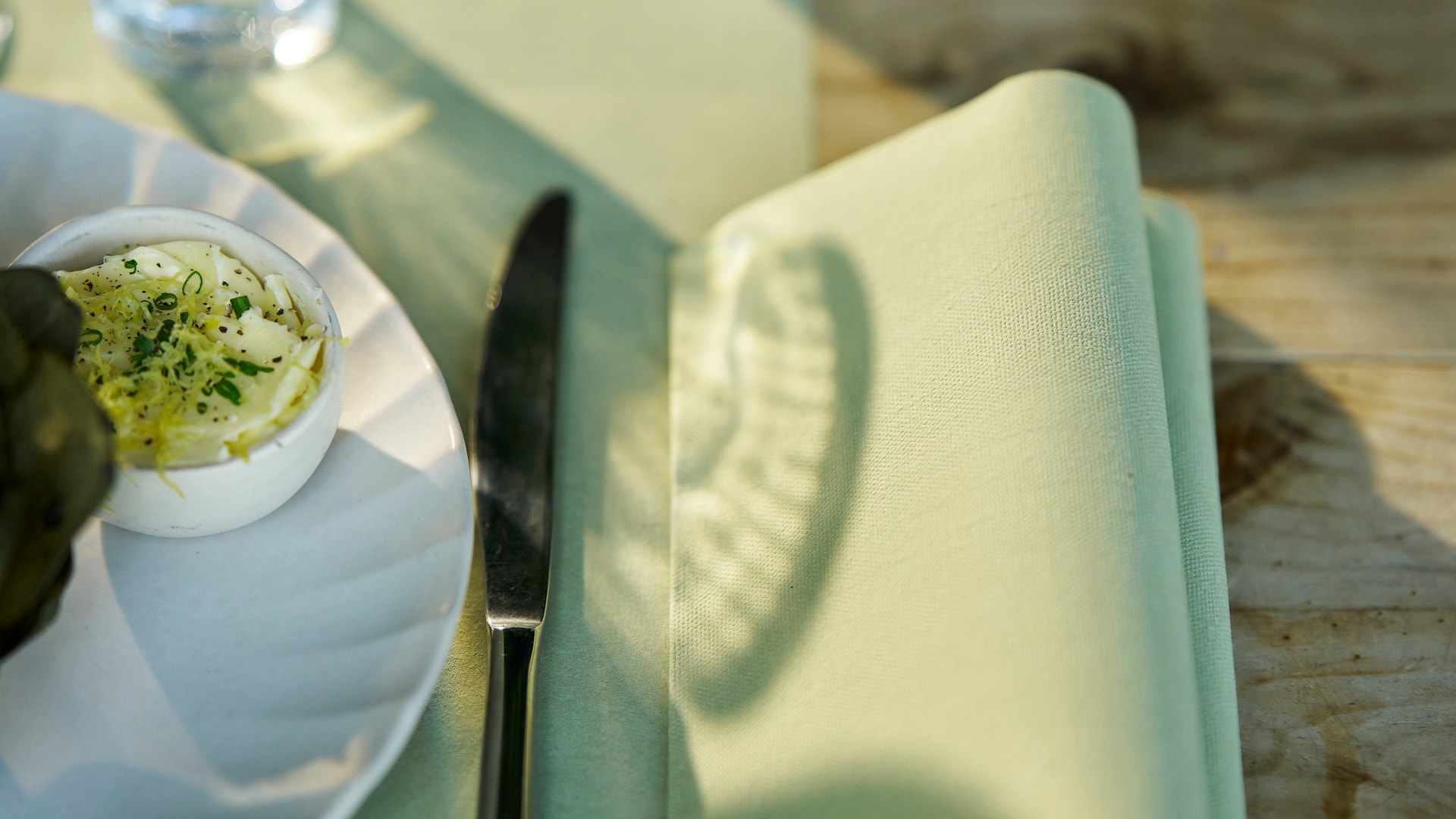 En bordsdukning med en pistaschgrön servett, vattenglas och tallrik med mat på ett trädgårdsbordet.