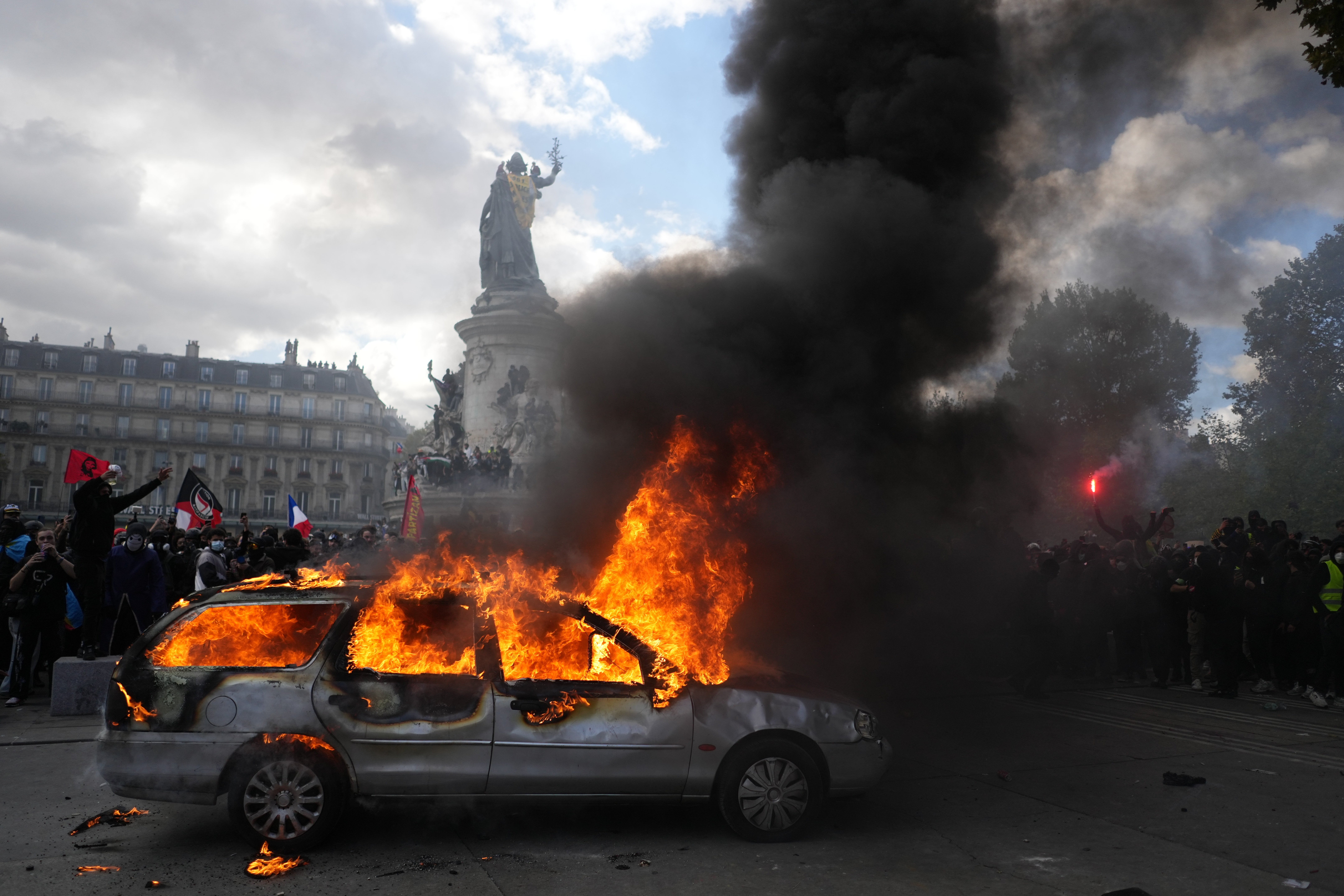 En bil brinner med höga lågor och svart rök på ett torg i Paris. Bakom bilen står en folkmassa framför en staty.