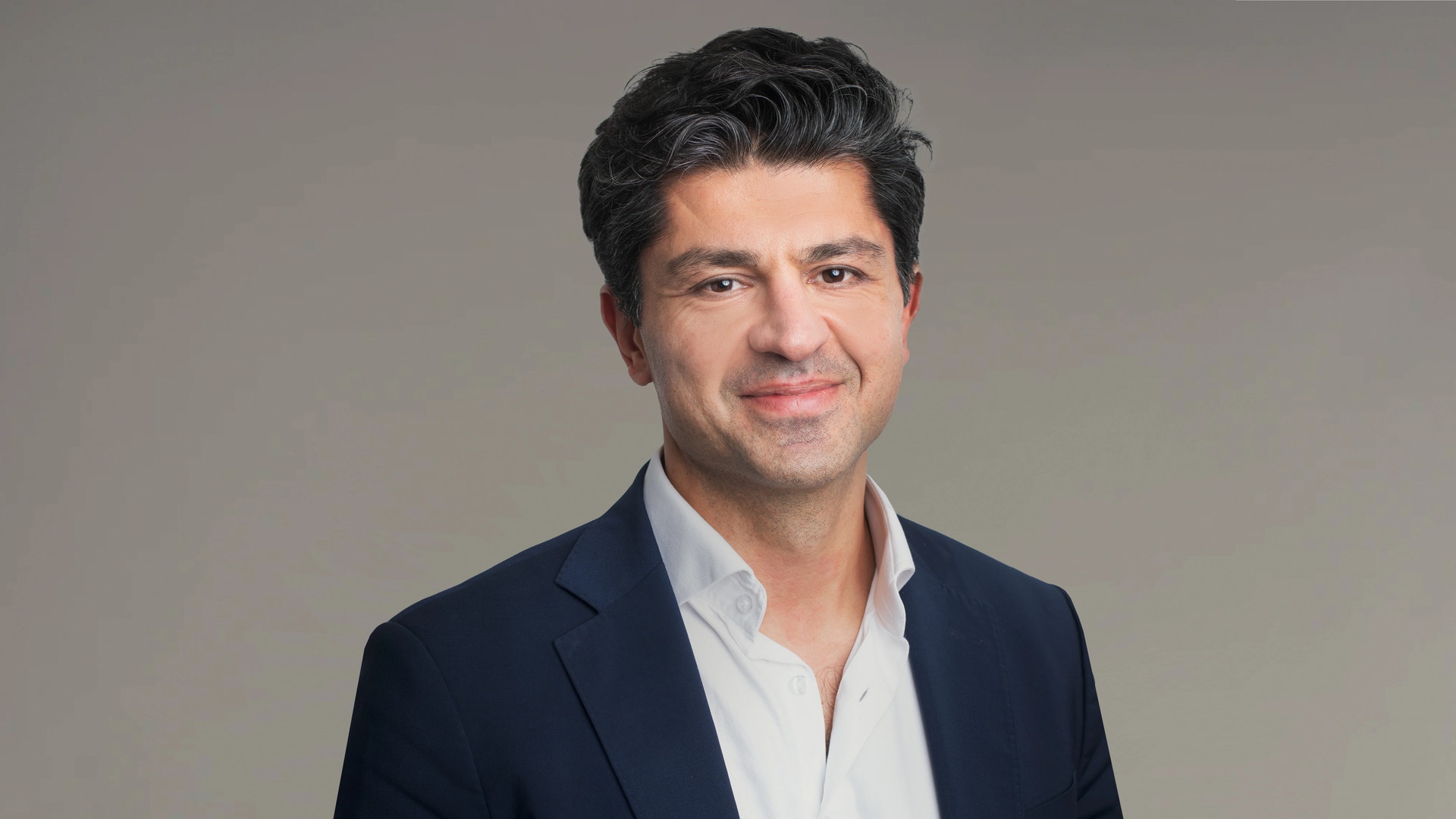 Porträttfoto av en man i mörkblå kavaj och vit skjorta mot neutral bakgrund.