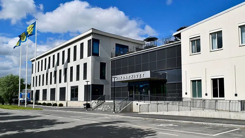 Exteriörbild av Riksarkivets byggnad i Täby, med svenska flaggor och en parkeringsplats i förgrunden.