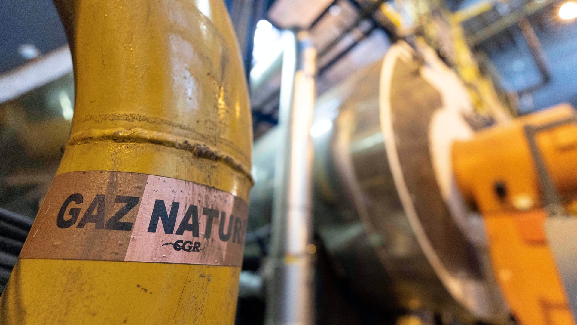 Närbild av en gul gaspipa märkt med etikett för naturgas, med industriell utrustning i bakgrunden.