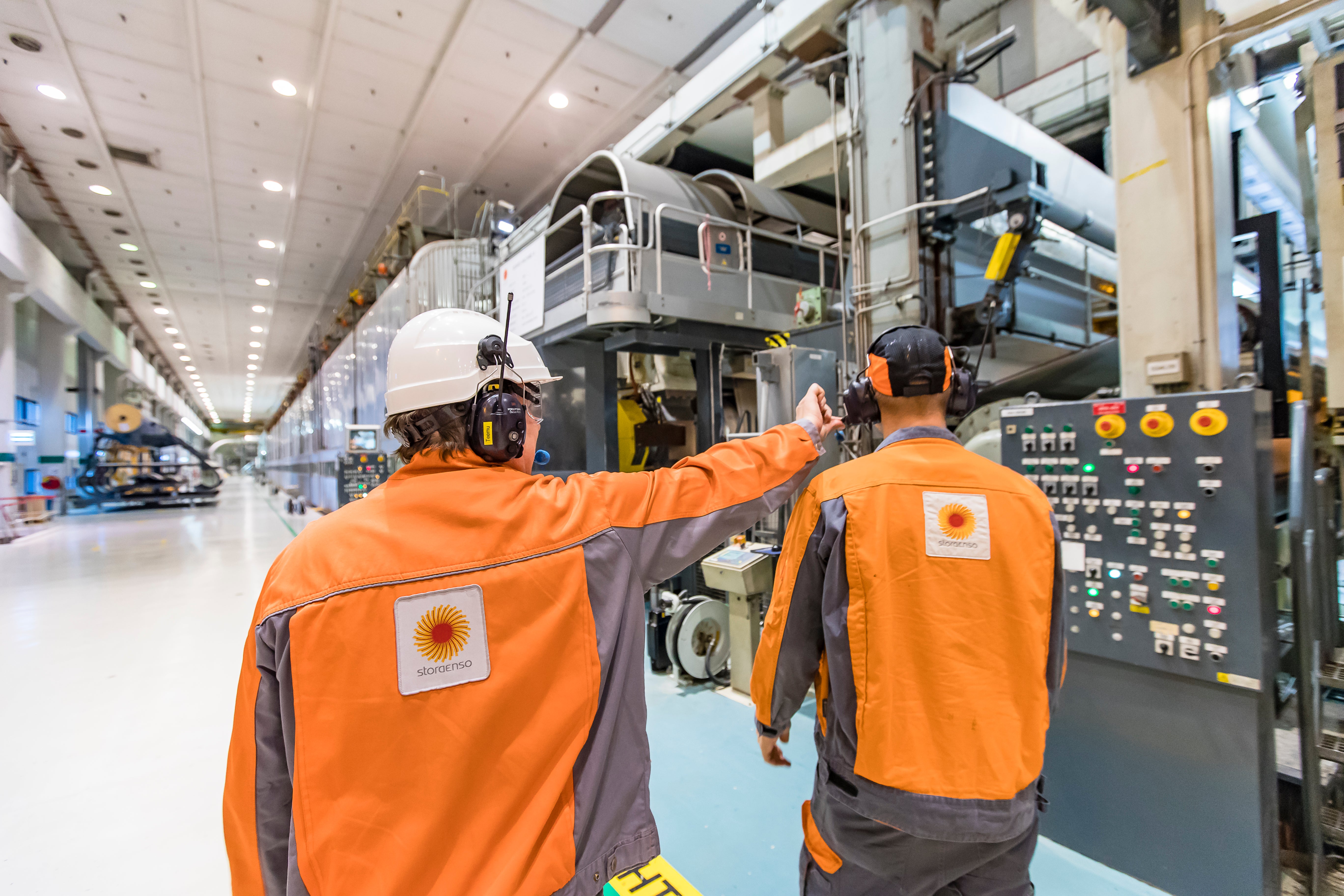 Två arbetare i orange säkerhetskläder och vita hjälmar arbetar vid en stor industriell maskin i en fabrikshall.