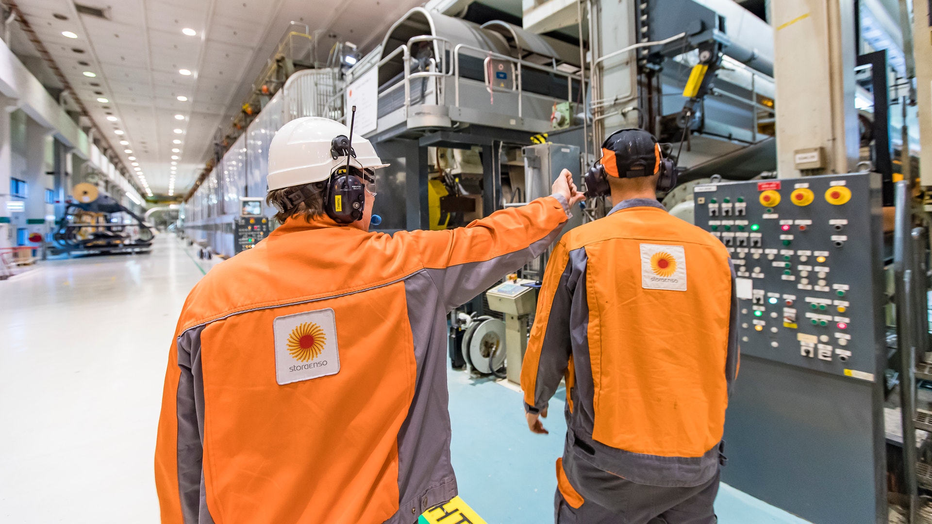 Två arbetare i orange säkerhetskläder och vita hjälmar arbetar vid en stor industriell maskin i en fabrikshall.