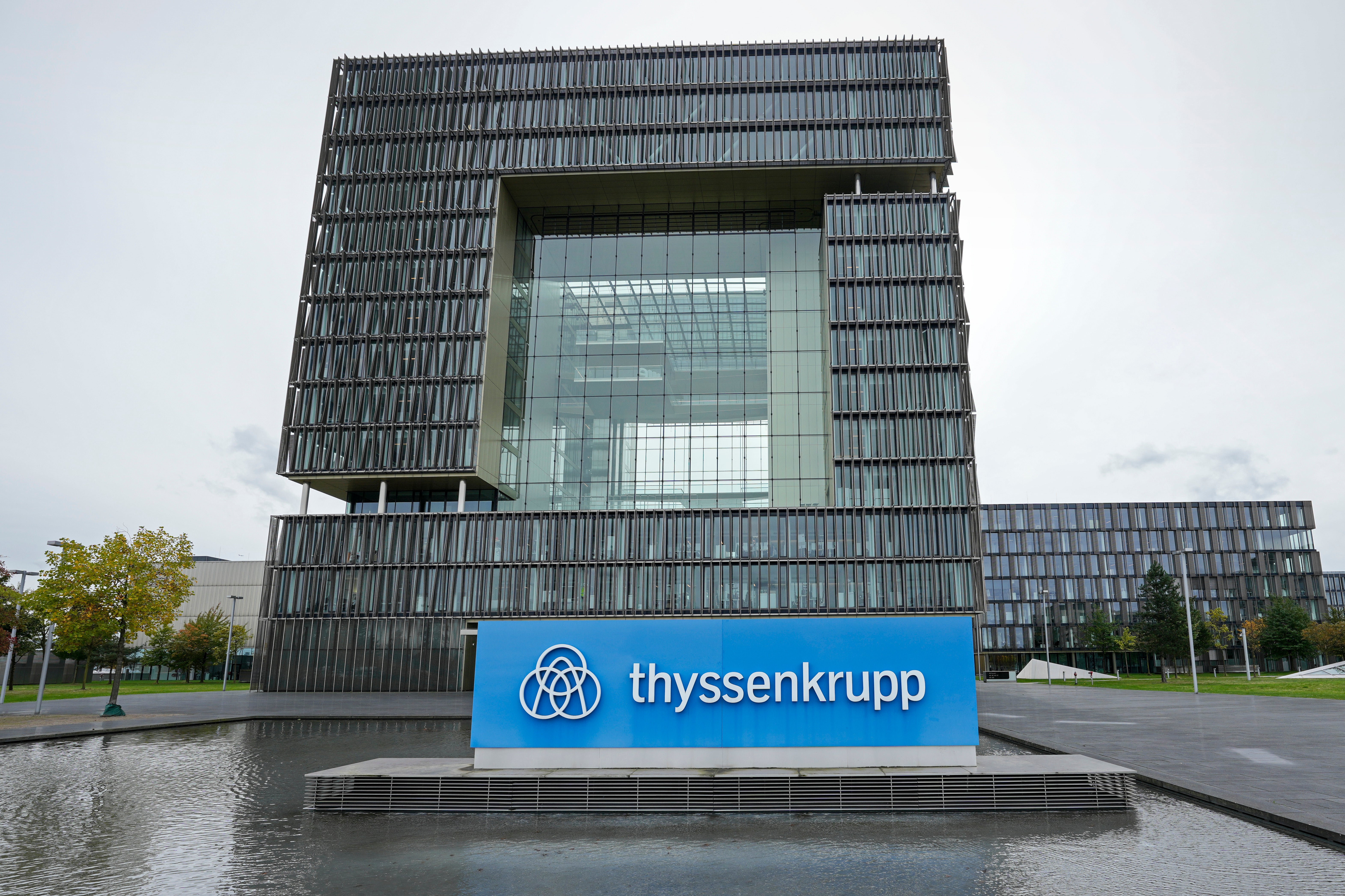 Thyssenkrupps moderna huvudkontor med karakteristisk arkitektur - en kvadratisk byggnad med mörkgrå fasad och stort mittfönster, omgiven av vatten och gröna områden. Blå skylt med Thyssenkrupps logotyp och företagsnamn i förgrunden.