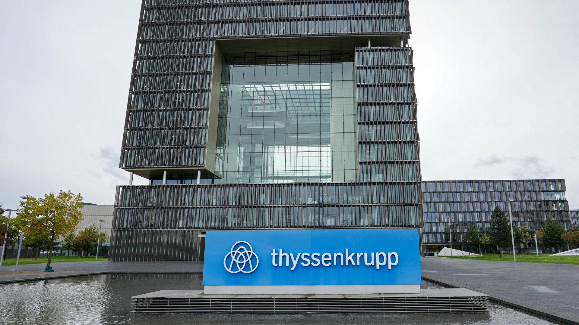 Thyssenkrupps moderna huvudkontor med karakteristisk arkitektur - en kvadratisk byggnad med mörkgrå fasad och stort mittfönster, omgiven av vatten och gröna områden. Blå skylt med Thyssenkrupps logotyp och företagsnamn i förgrunden.