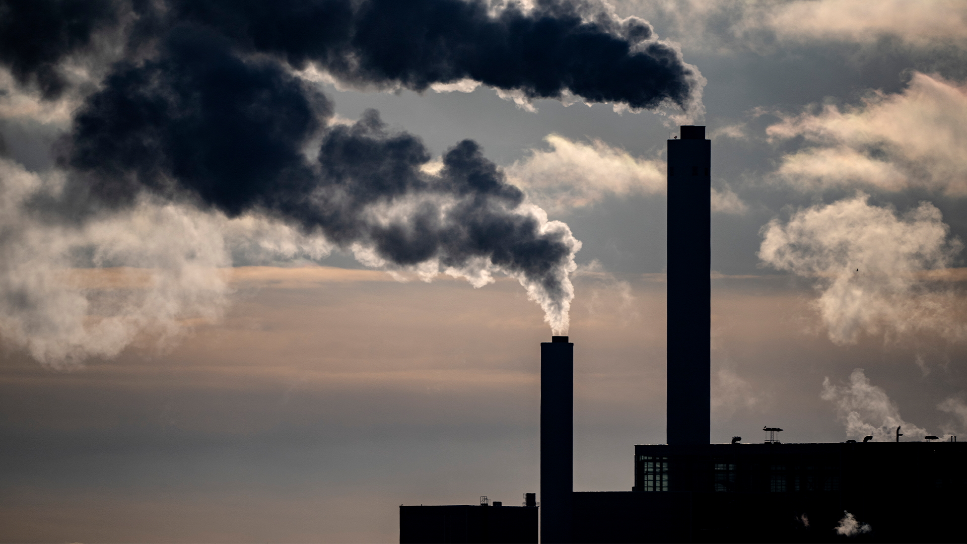 Två höga industriella skorstenar som släpper ut tjock mörk rök och vit ånga mot en molnig himmel. Bilden illustrerar miljöföroreningar och utsläpp från industriell verksamhet.