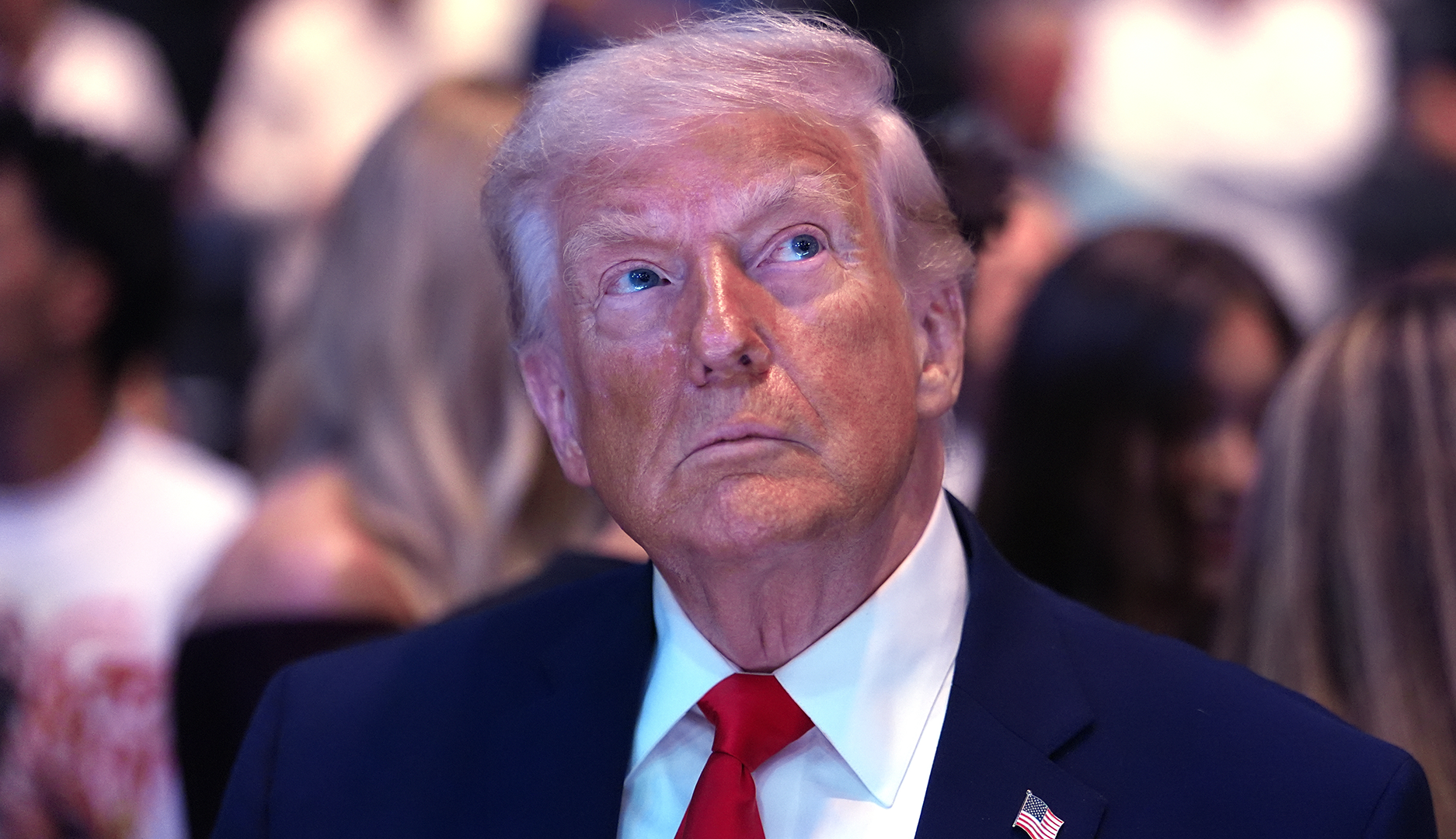 Porträtt av en man i mörkblå kostym och röd slips framför en suddig bakgrund med människor.