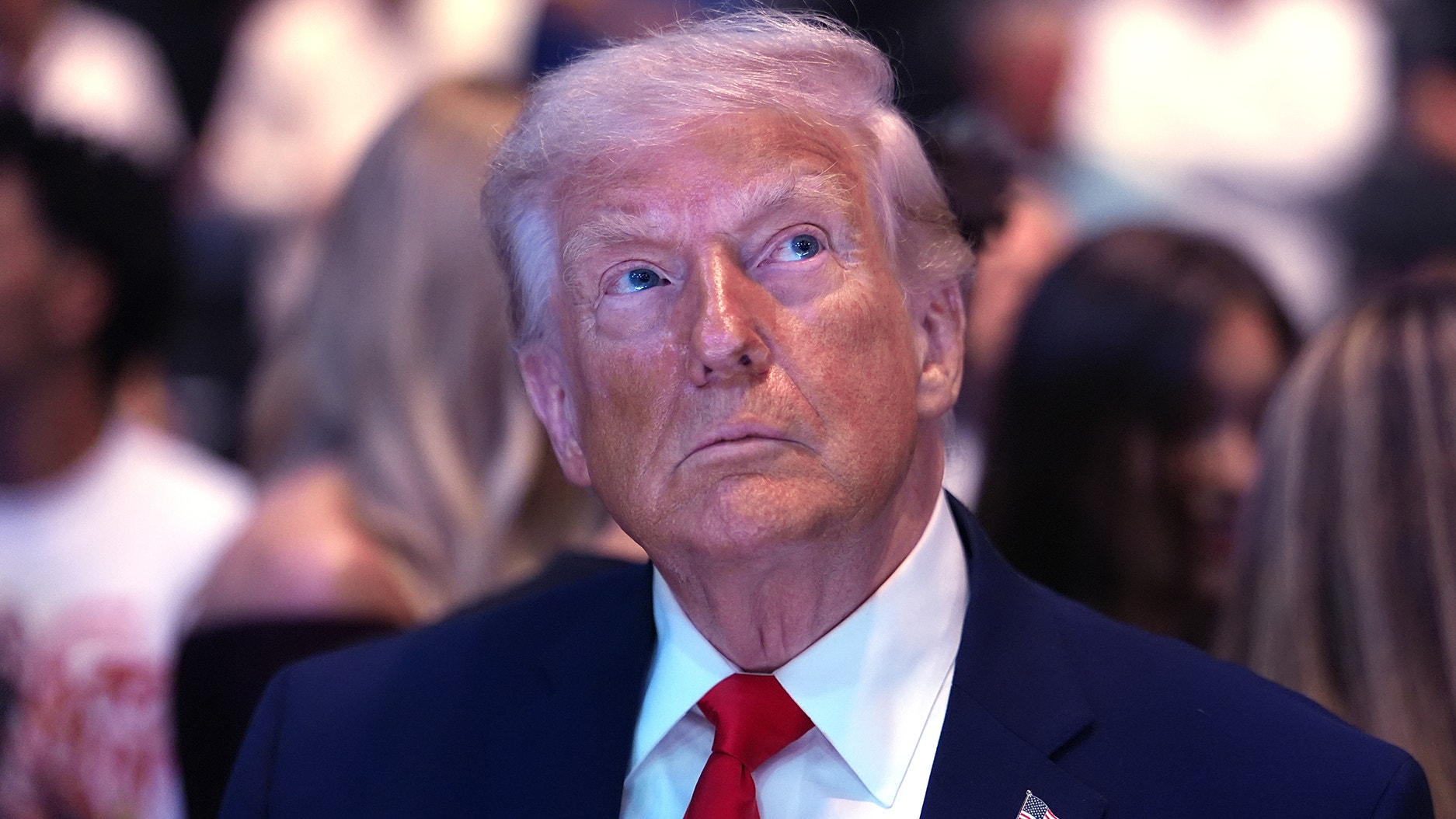 Porträtt av en man i mörkblå kostym och röd slips framför en suddig bakgrund med människor.