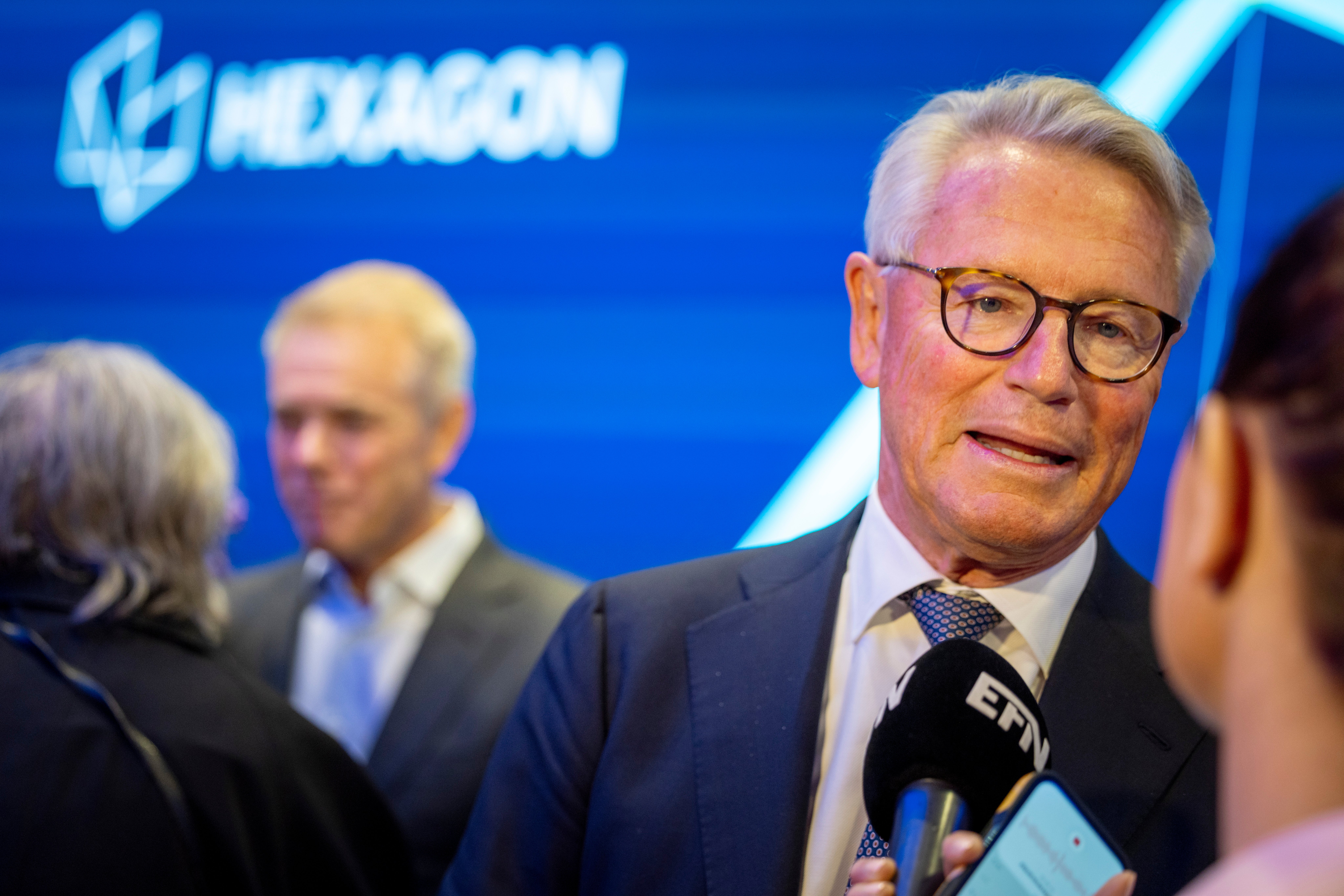 En man i glasögon och mörk kostym blir intervjuad med mikrofon vid en professionell event med blå bakgrund.