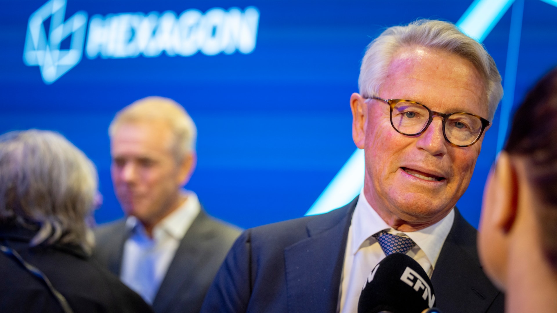 En man i glasögon och mörk kostym blir intervjuad med mikrofon vid en professionell event med blå bakgrund.