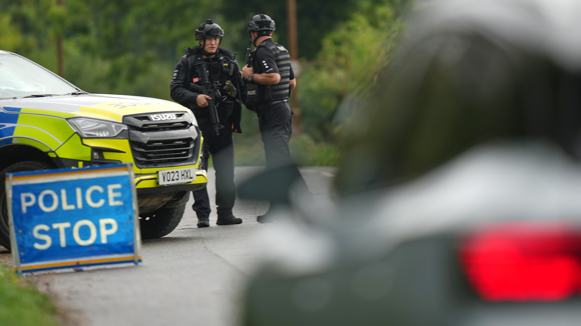 Beväpnade poliser står vid en polisbil och en avstängning med skylten "Police Stop" på en väg.