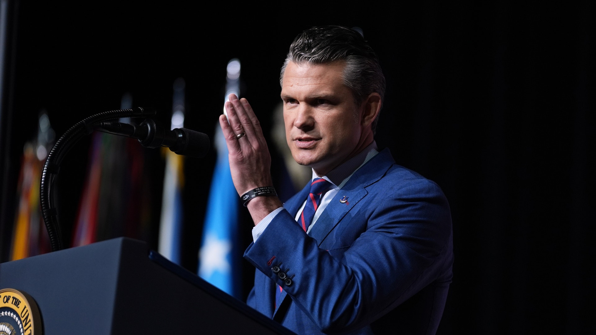 Man i blå kostym talar vid ett podium med presidentens sigill framför en svart bakgrund med flaggor.