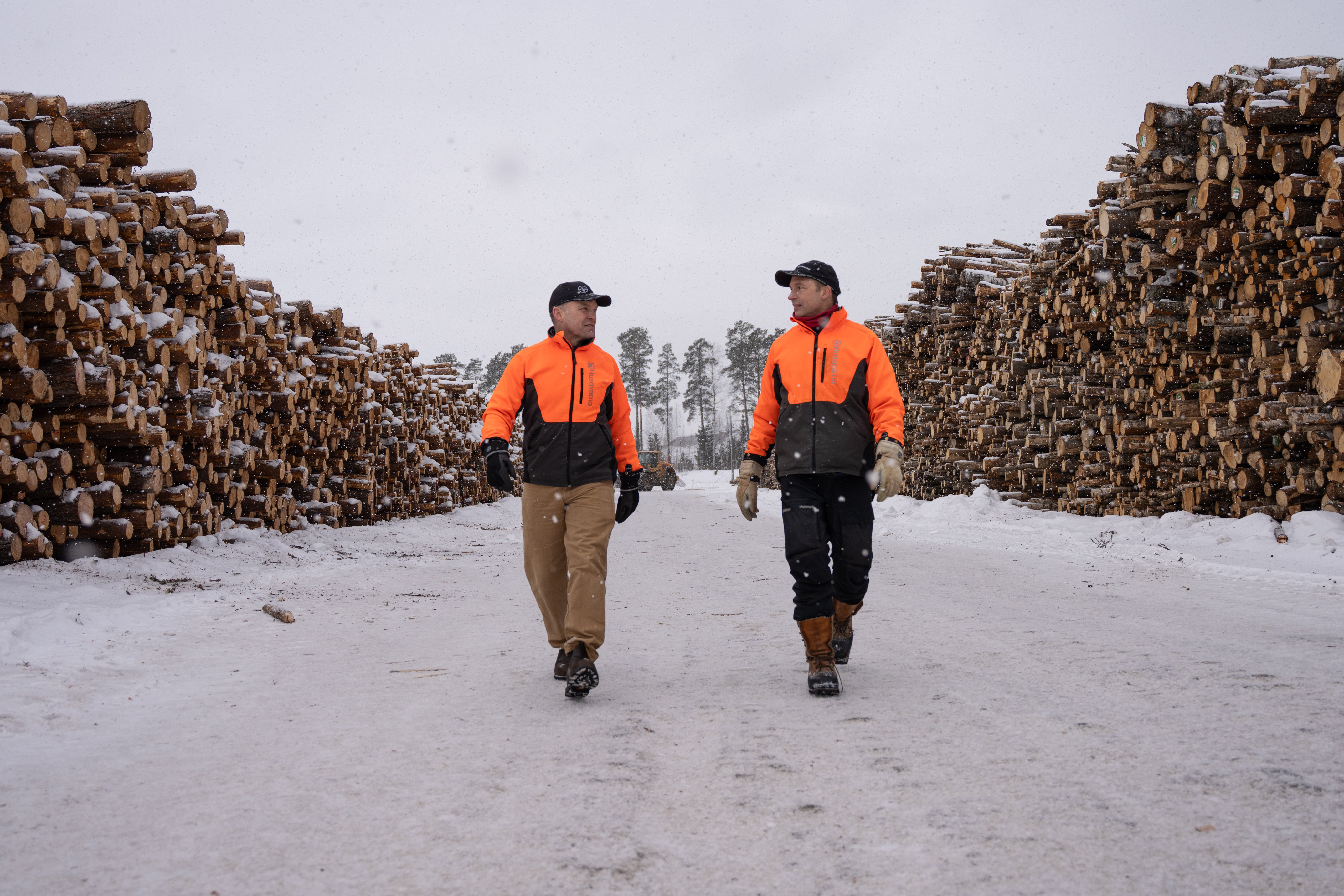 Två arbetare i orange västar går mellan höga stockar vid en timmerterminalen under vinterklimat.