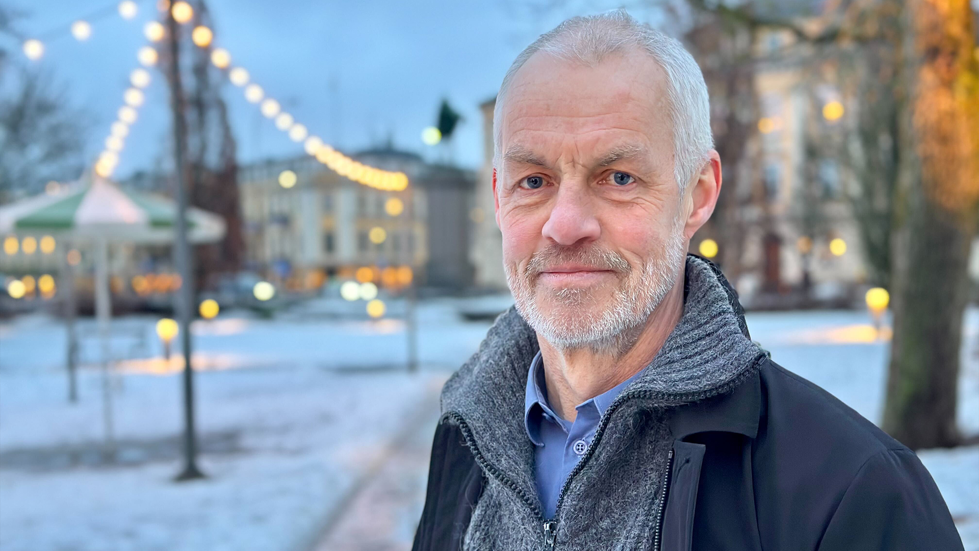 Porträtt av en äldre man i vintermiljö med belyst stadstorg i bakgrunden