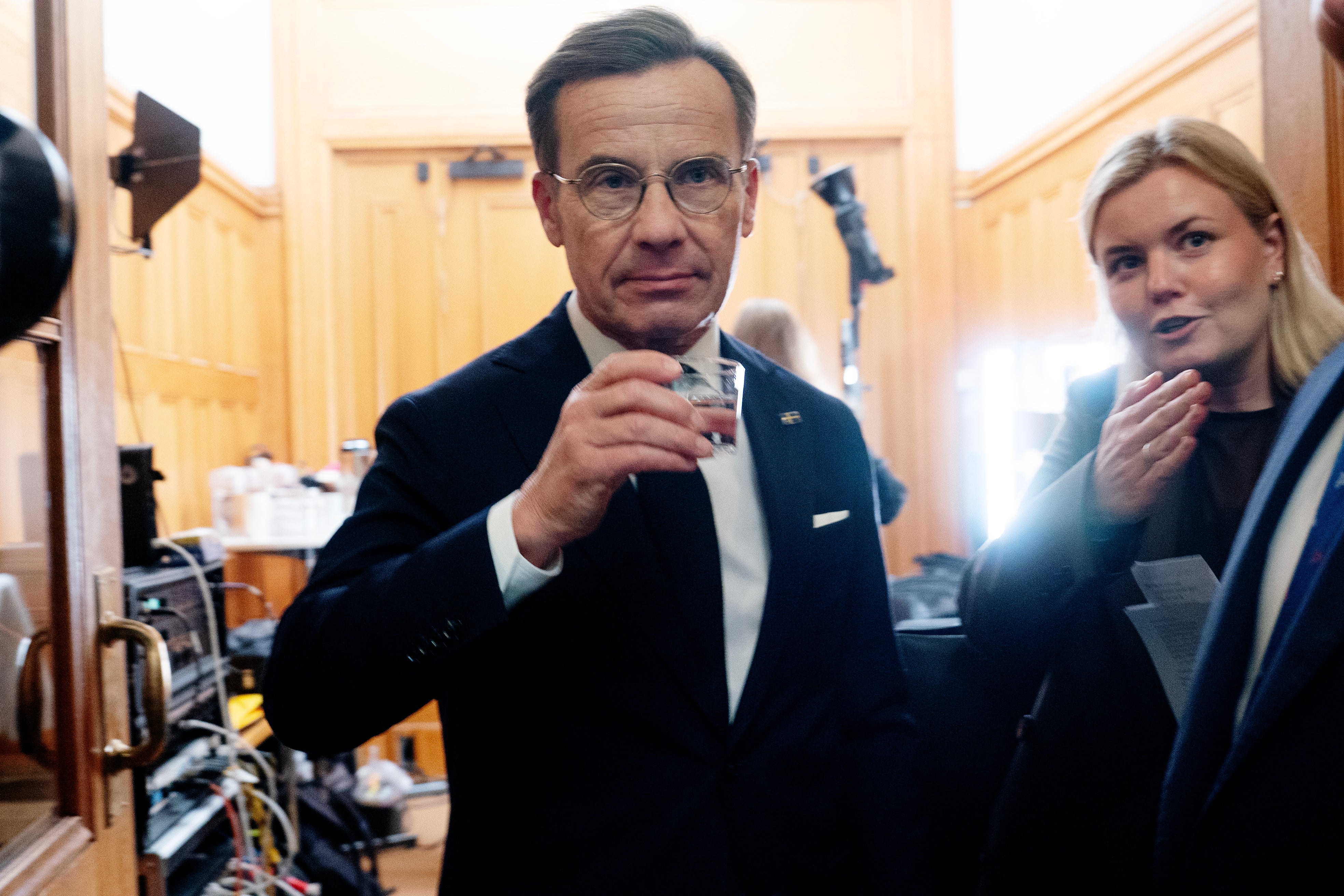 Man i kostym dricker ur ett glas i en träpanelerad korridor, med en kvinna och kamerautrustning i bakgrunden.