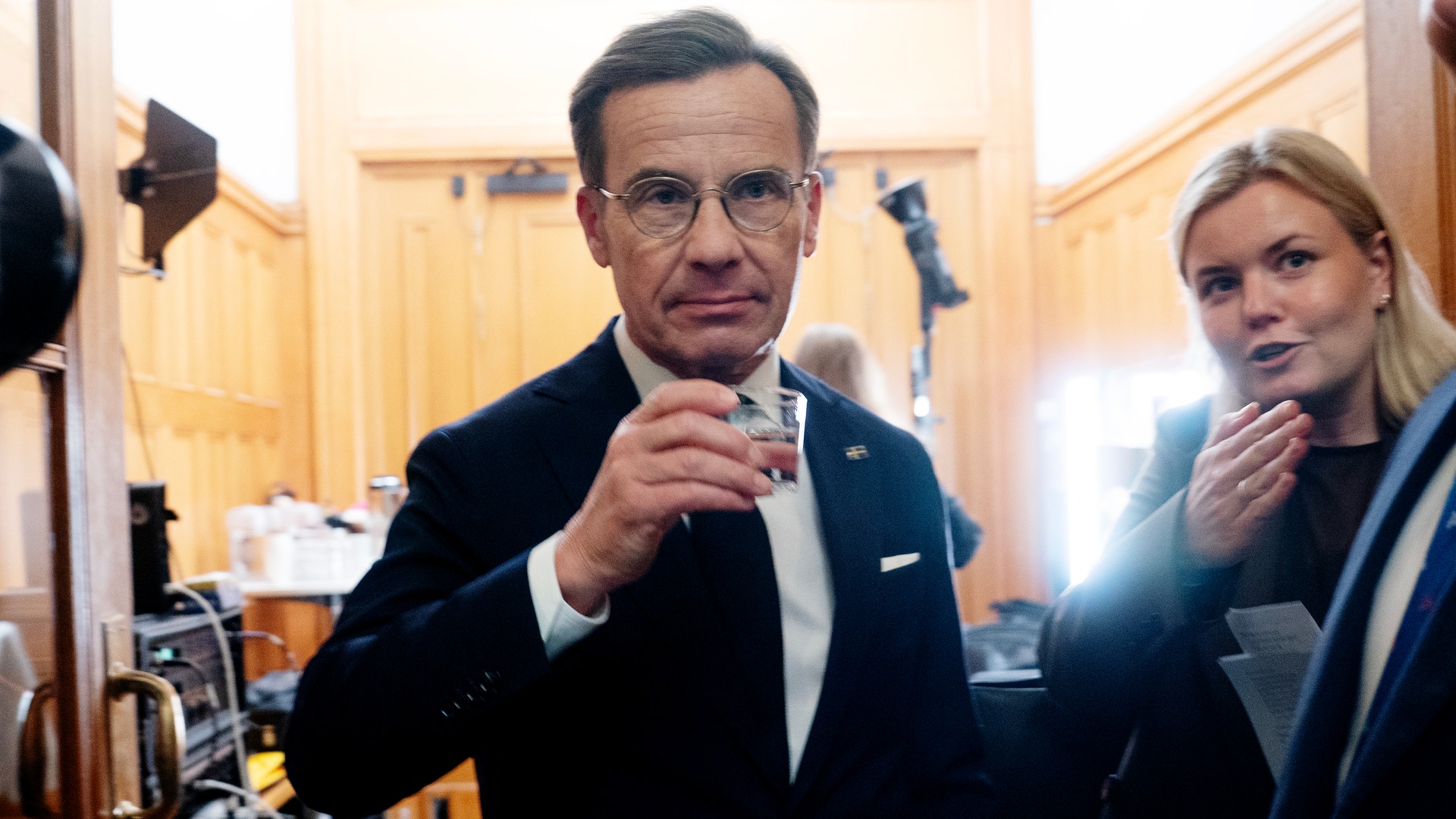 Man i kostym dricker ur ett glas i en träpanelerad korridor, med en kvinna och kamerautrustning i bakgrunden.