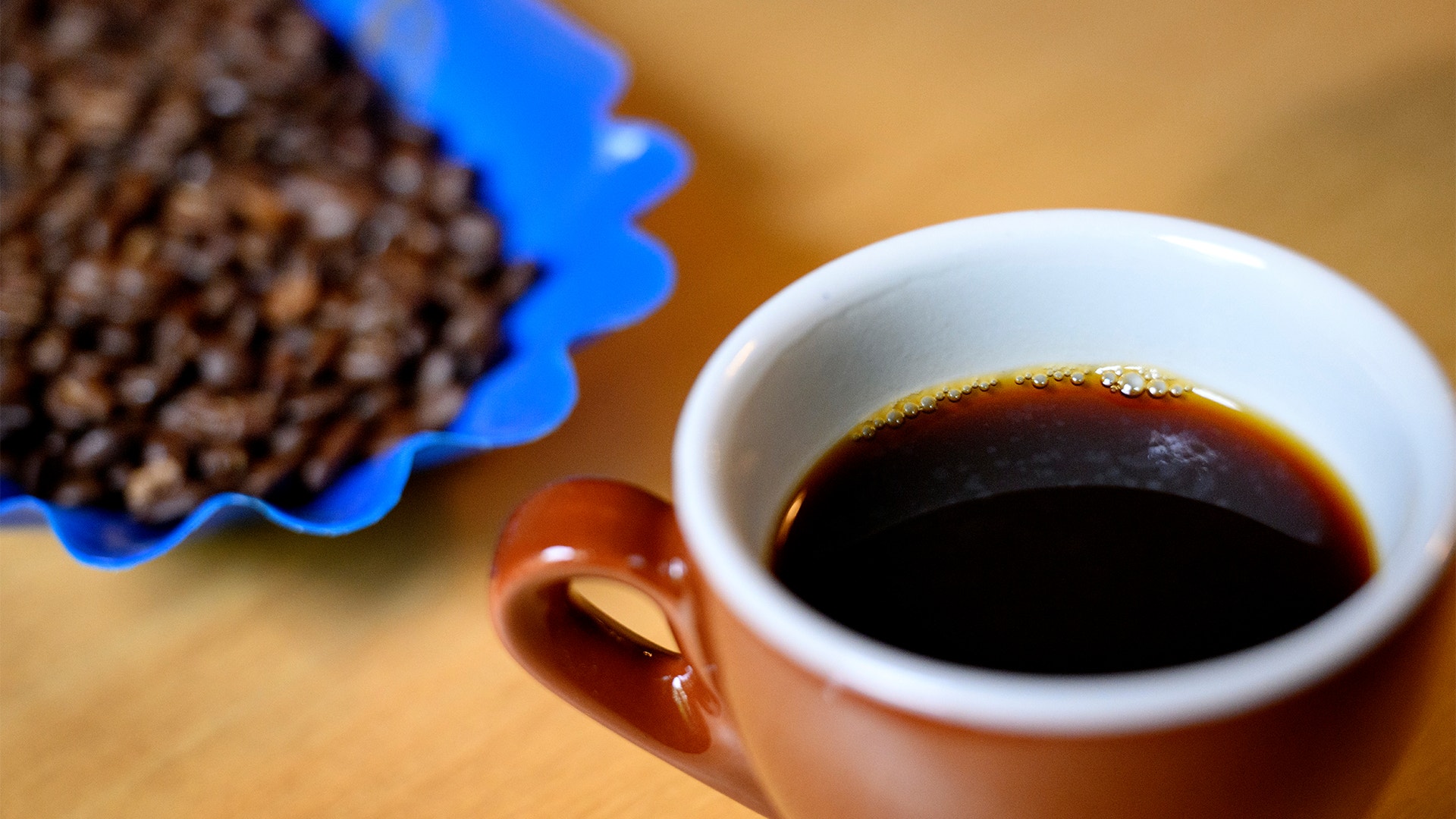 En kopp med svart kaffe bredvid en blå behållare fylld med kaffebönor på en ljus träbakgrund.